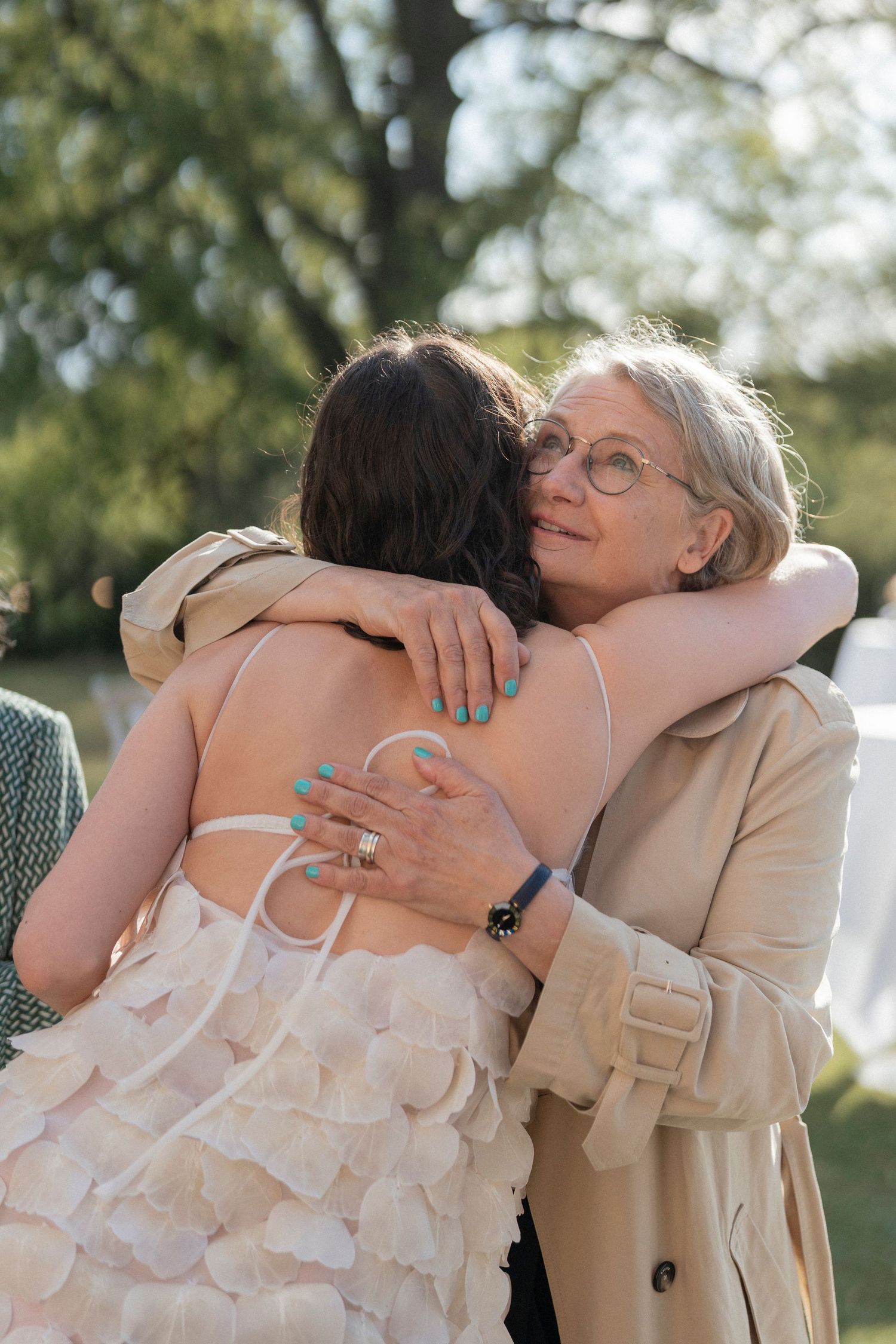 A bride in a white petal-layered wedding dress embraces an older woman in glasses and a beige trench coat outdoors.