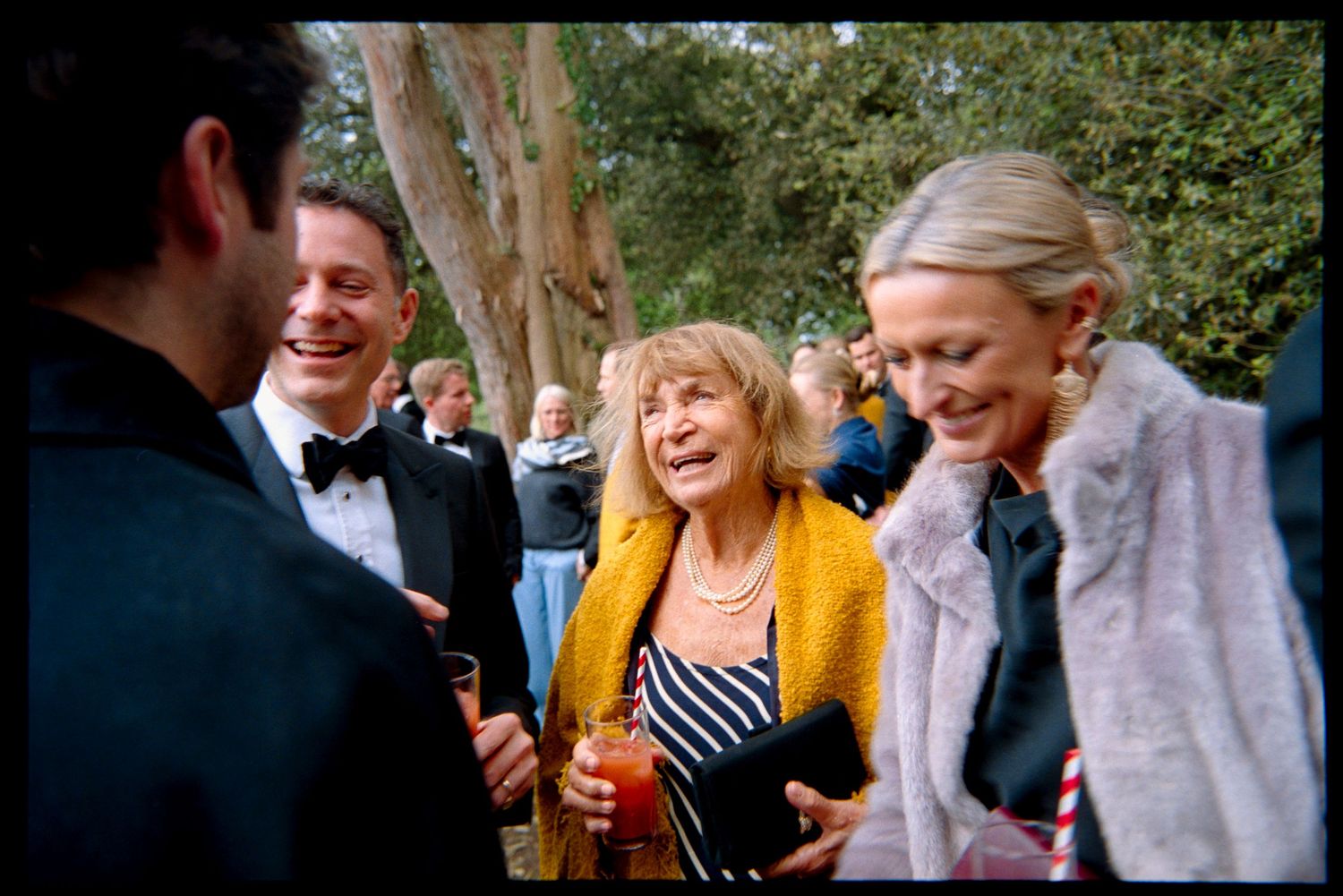 Smiling wedding guests chat and laugh outdoors under trees, a woman in a yellow cardigan holding a drink.
