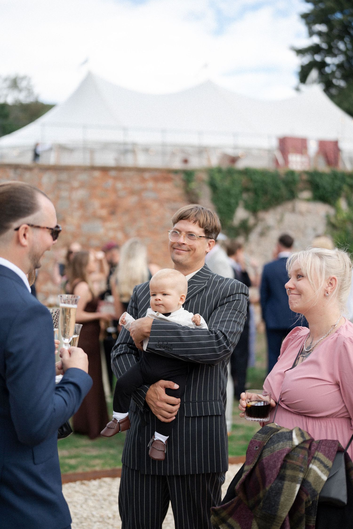 Man in pinstripe suit holding baby chats with guests at an outdoor wedding reception near a white tent.