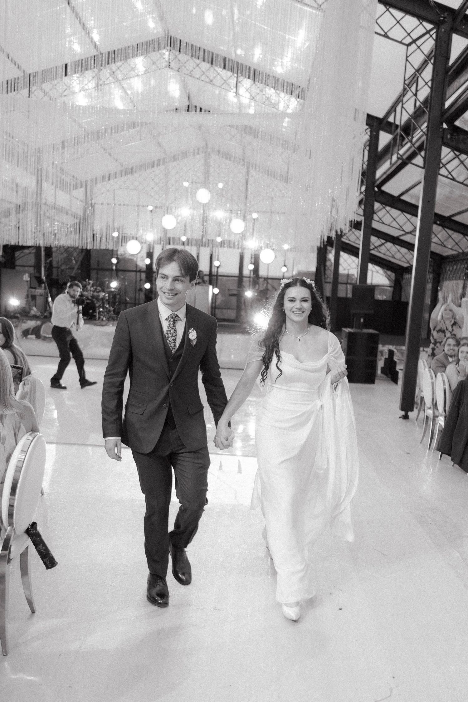 Bride and groom walk hand-in-hand in a decorated wedding venue in a black and white photo.