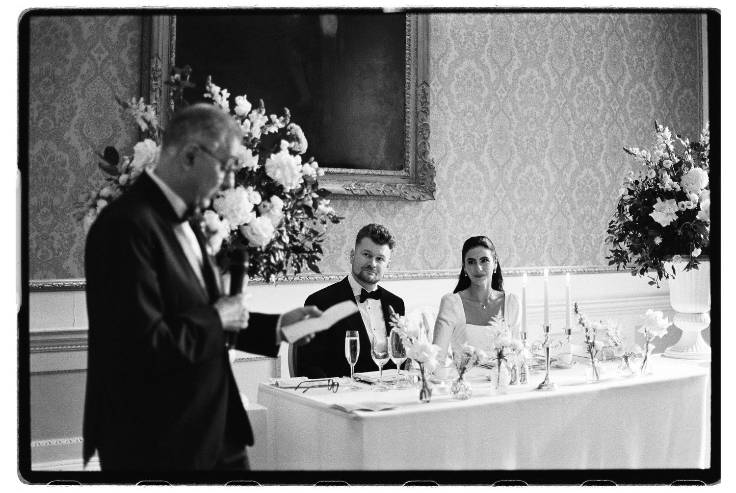 Black and white wedding photo of a man giving a toast beside a seated bride and groom at an elegant head table with flowers and candles.