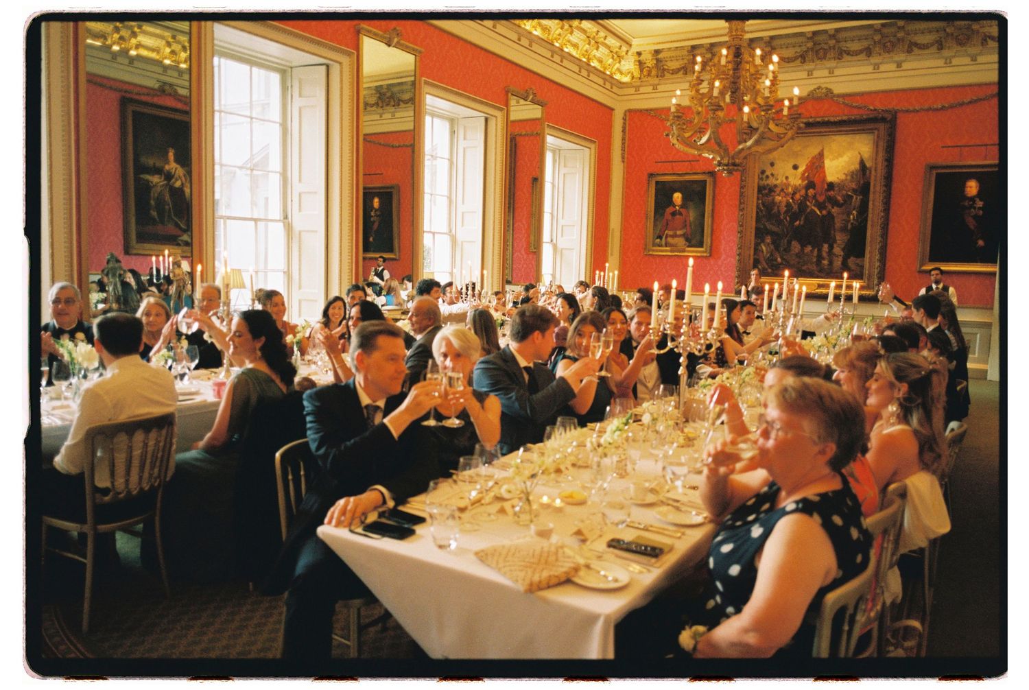 Elegant formal dinner party in a grand red-walled dining room with chandelier, guests seated at long tables with candles.