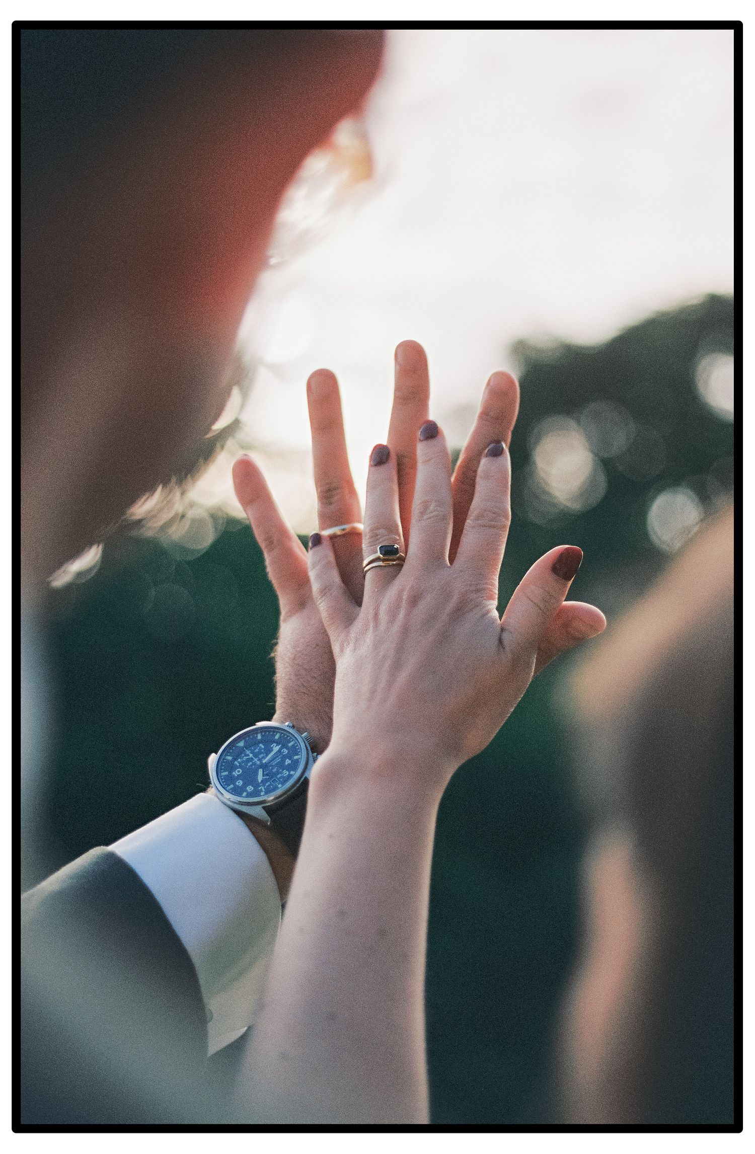 Two hands reaching towards each other showing wedding rings with a blue watch visible against a blurred background.