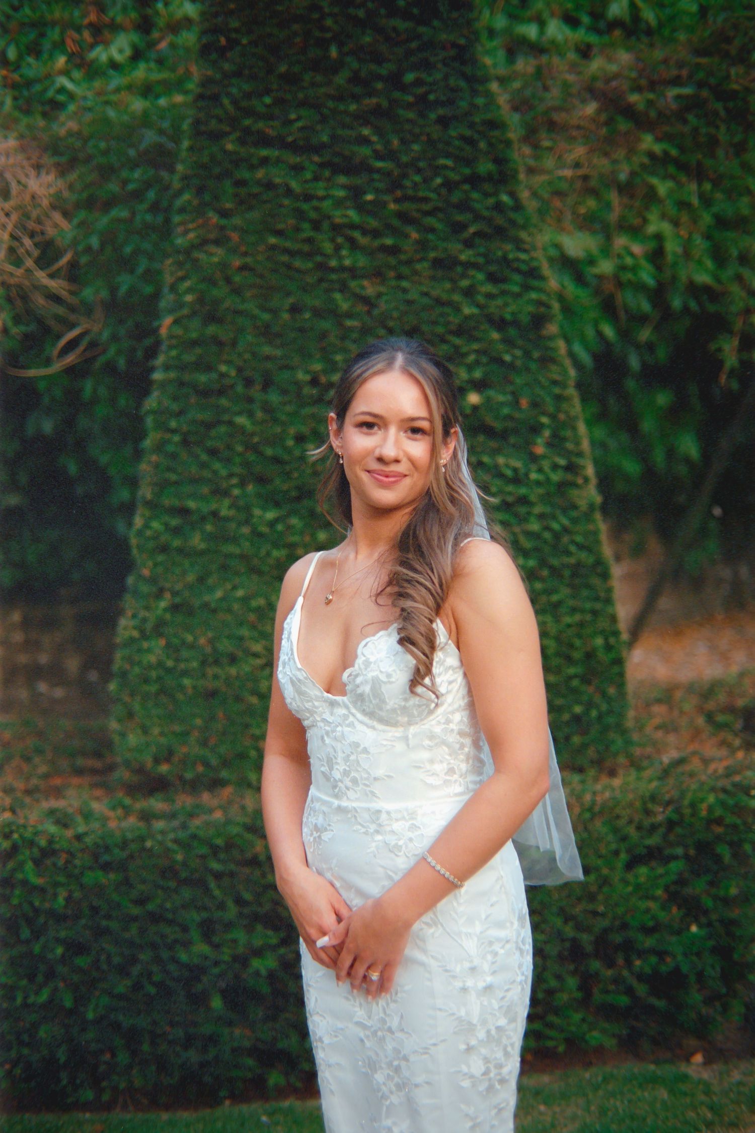 A bride in a white lace wedding dress poses outdoors against a green ivy-covered wall background.