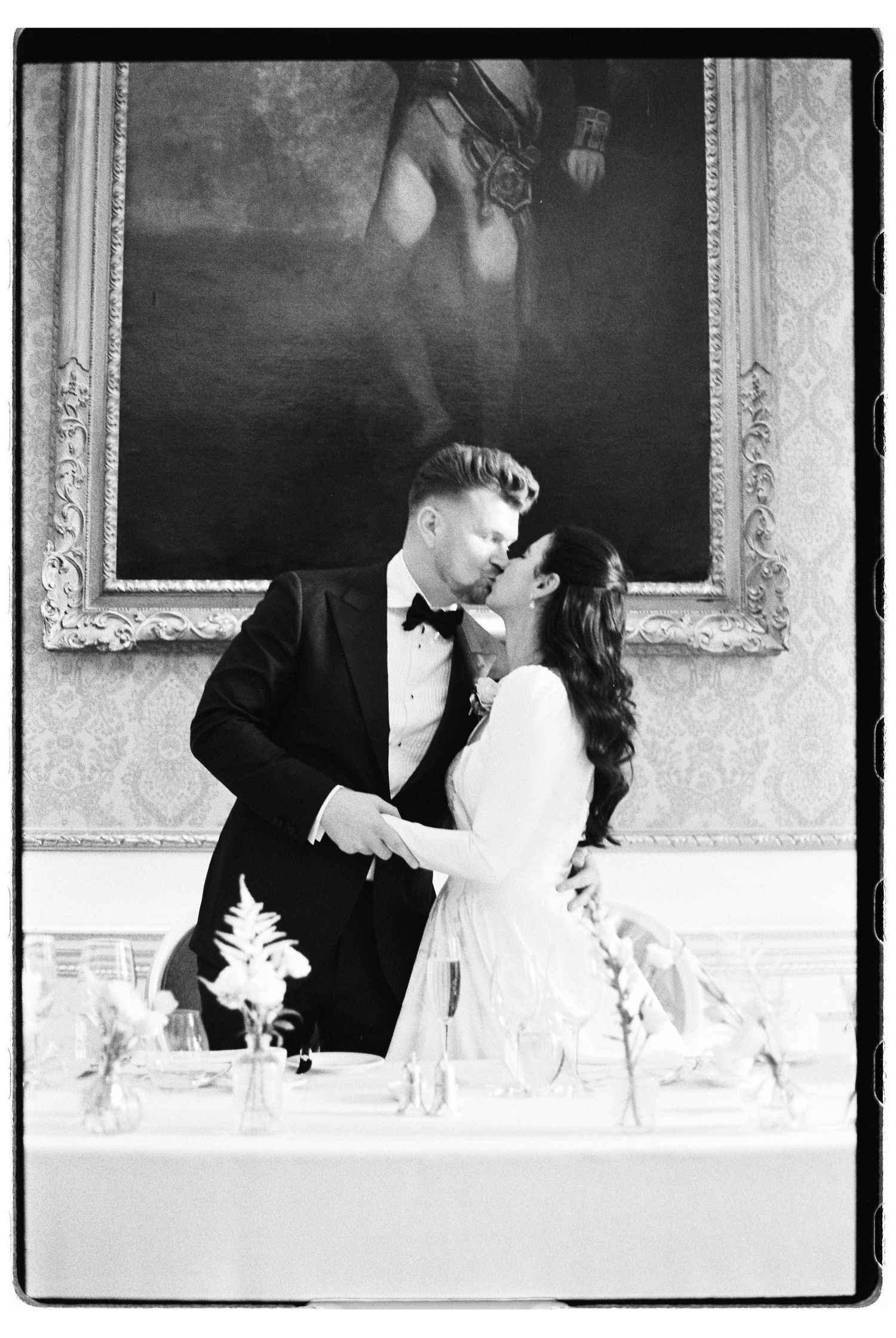 Black and white wedding photo of couple sharing a kiss at reception table with ornate painting backdrop.