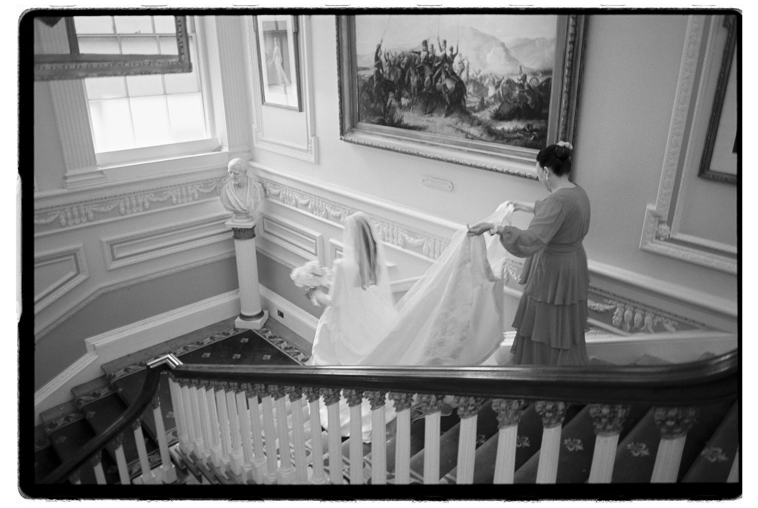 Black and white photo of a bride in a wedding gown with veil being adjusted on an elegant staircase.