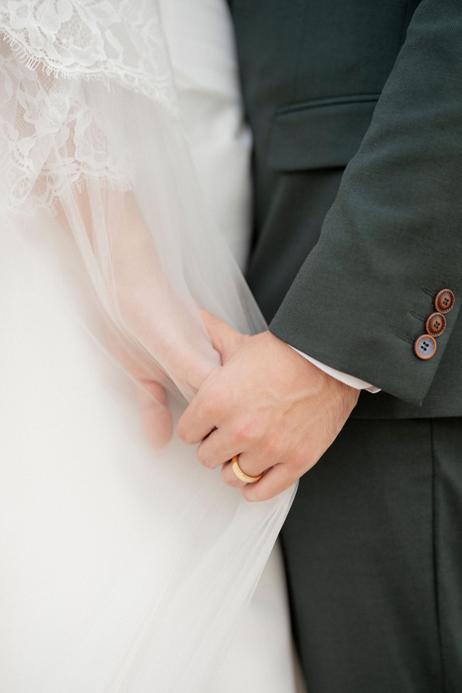 Close-up of newlyweds holding hands, groom wearing gold wedding band, bride in lace dress with veil.