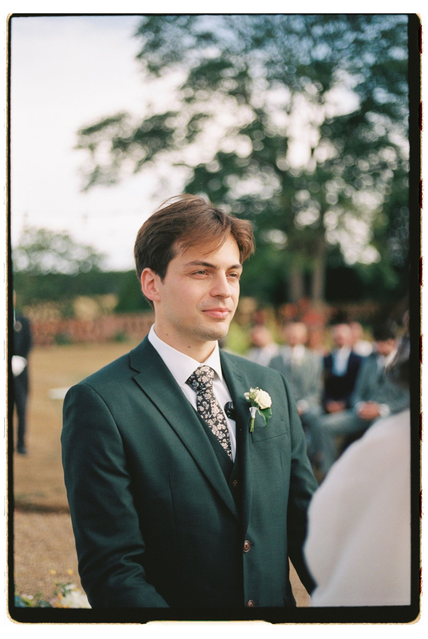 Groom in dark green suit with floral tie and boutonniere smiling at outdoor wedding ceremony.