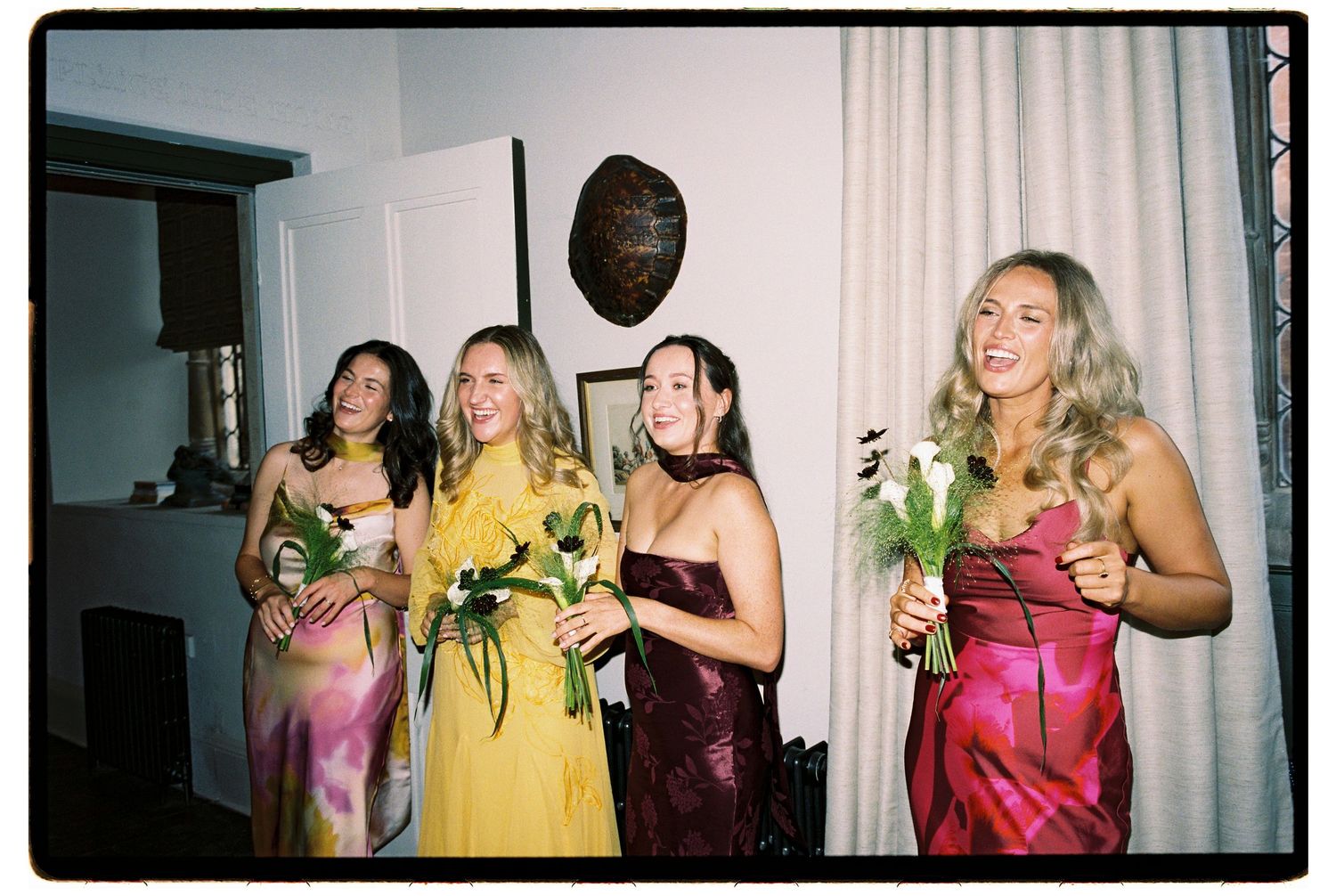 Four bridesmaids in colorful satin dresses holding white flowers pose together at a wedding reception.