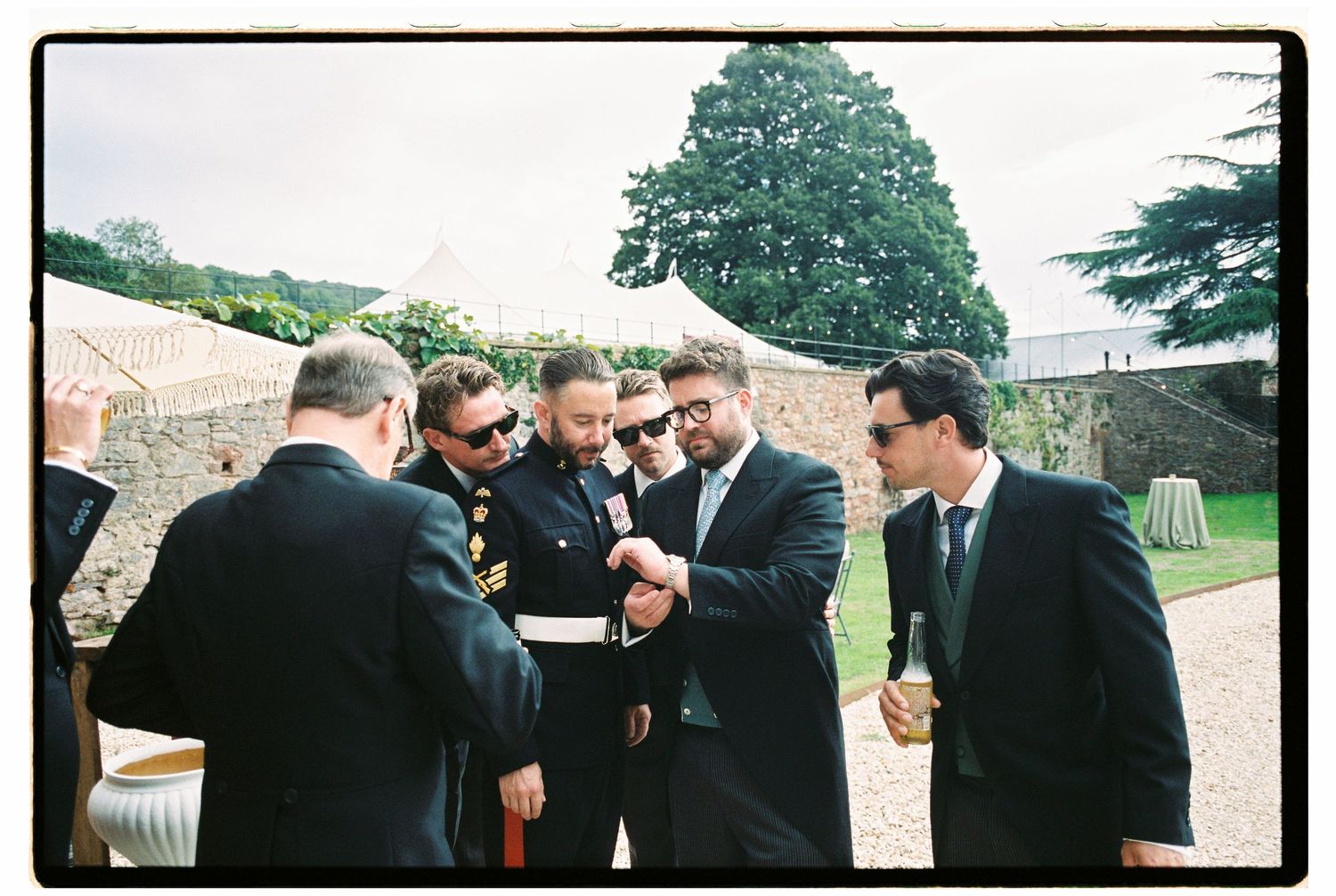 Groomsmen in suits gather outdoors at a wedding venue, examining details together near a stone wall.