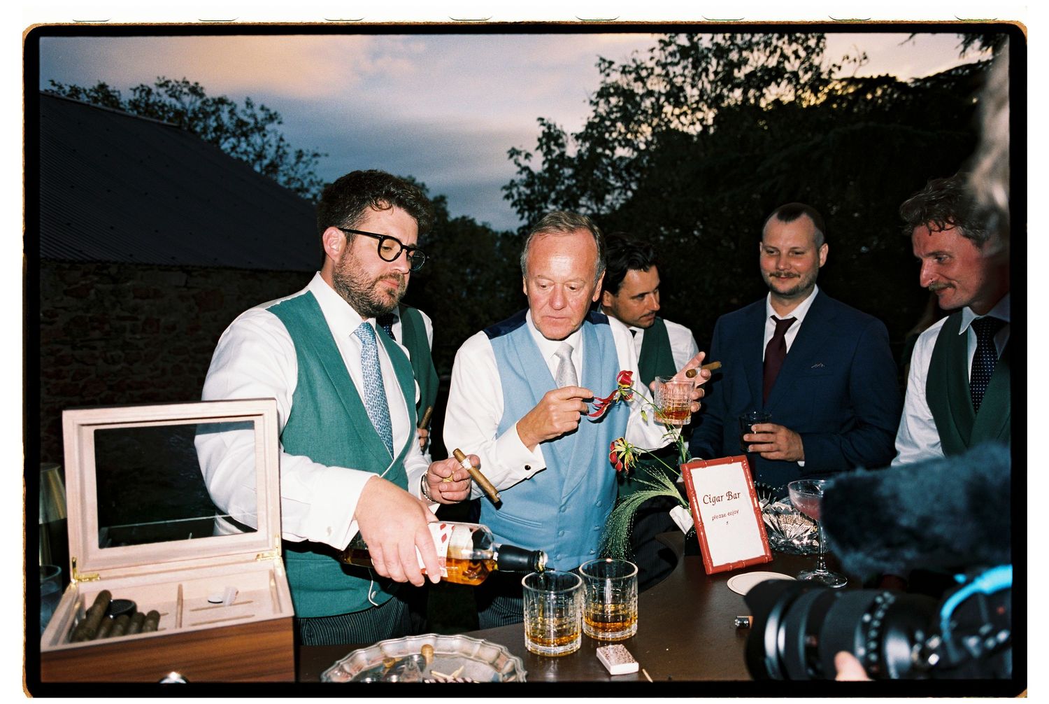 Groom in green vest pours whiskey at outdoor bar with guests during evening wedding reception.