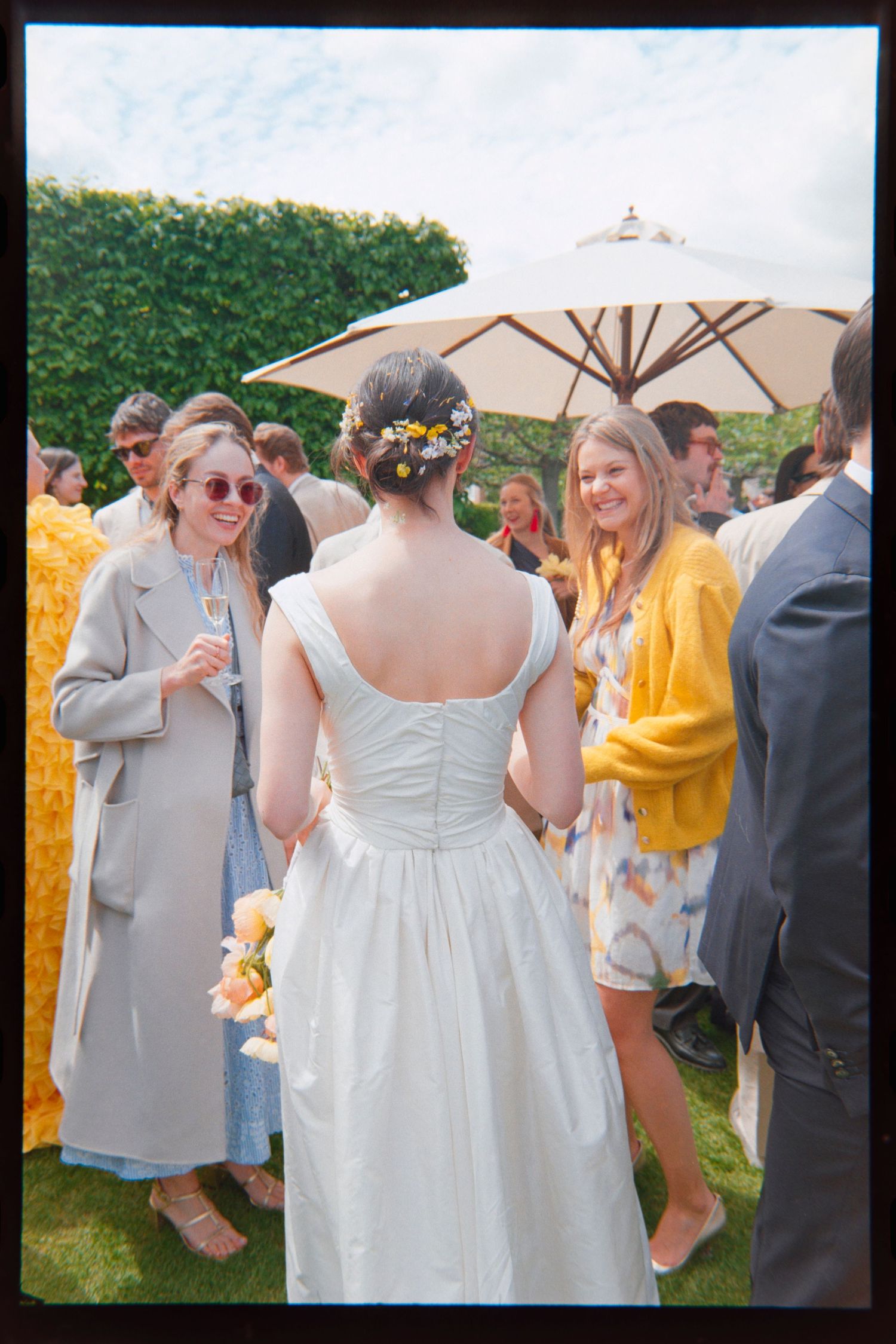 Bride in a white gown with a flower hair accessory chats with smiling wedding guests on a sunny lawn beneath a large patio umbrella.