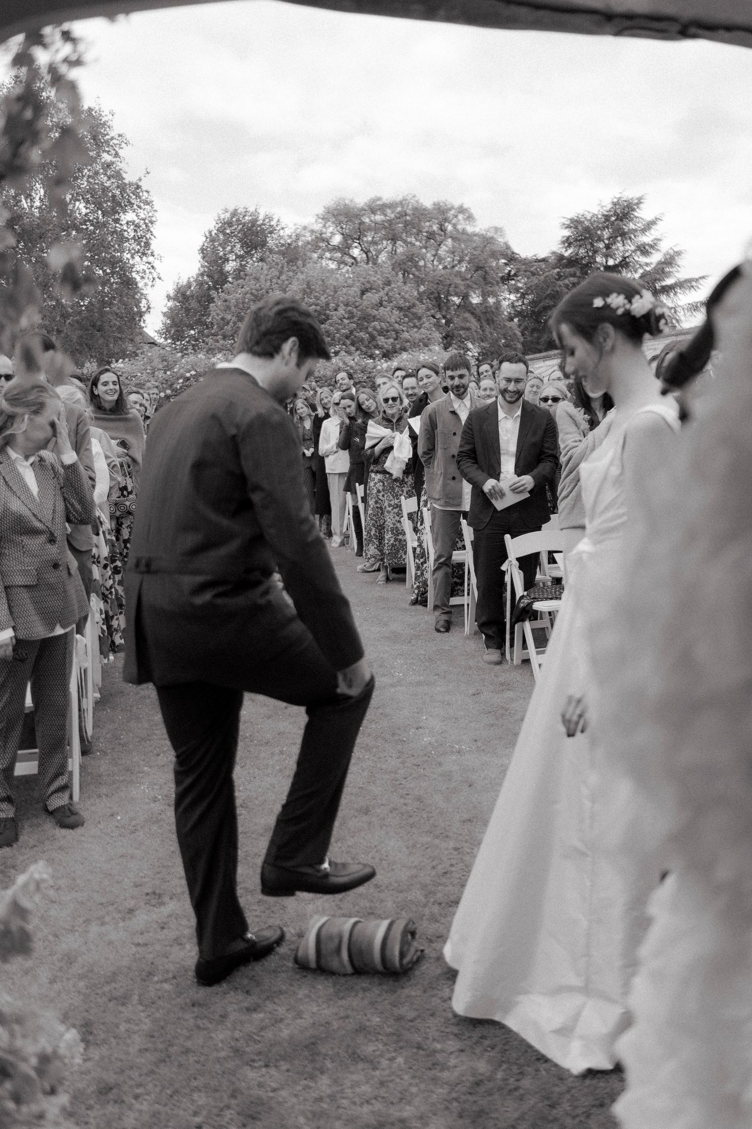 Groom stepping on a wrapped glass during an outdoor wedding ceremony as seated guests watch the bride in a white gown.