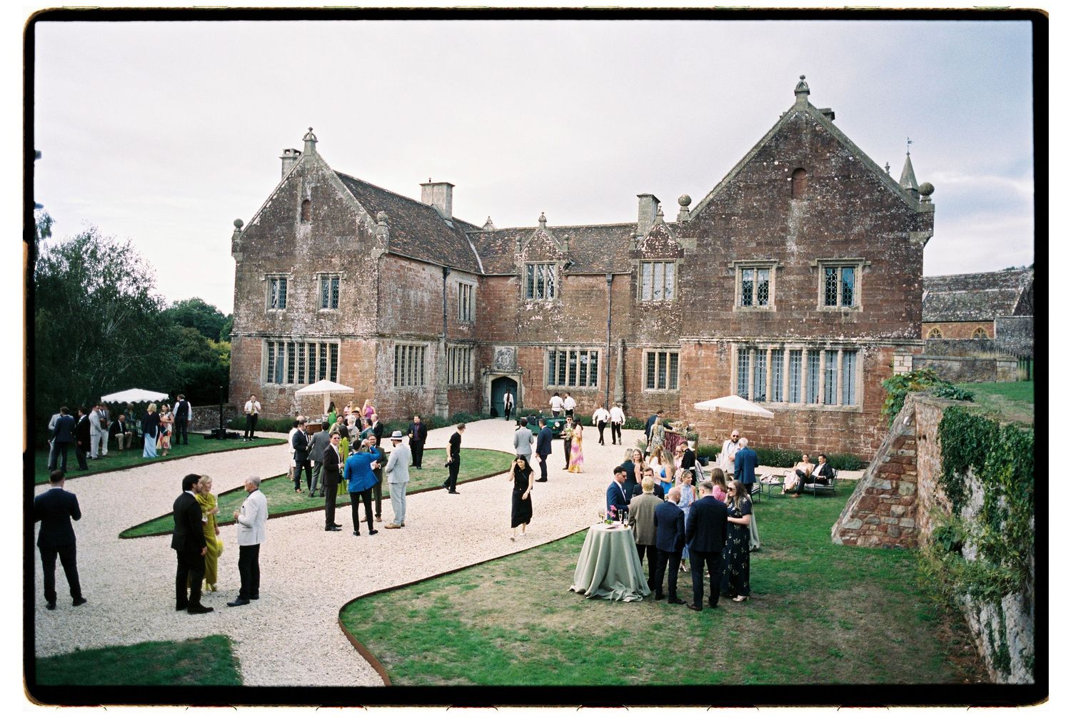 Wedding guests gather on a lawn and gravel path in front of a historic stone manor house with gabled roofs and mullioned windows.