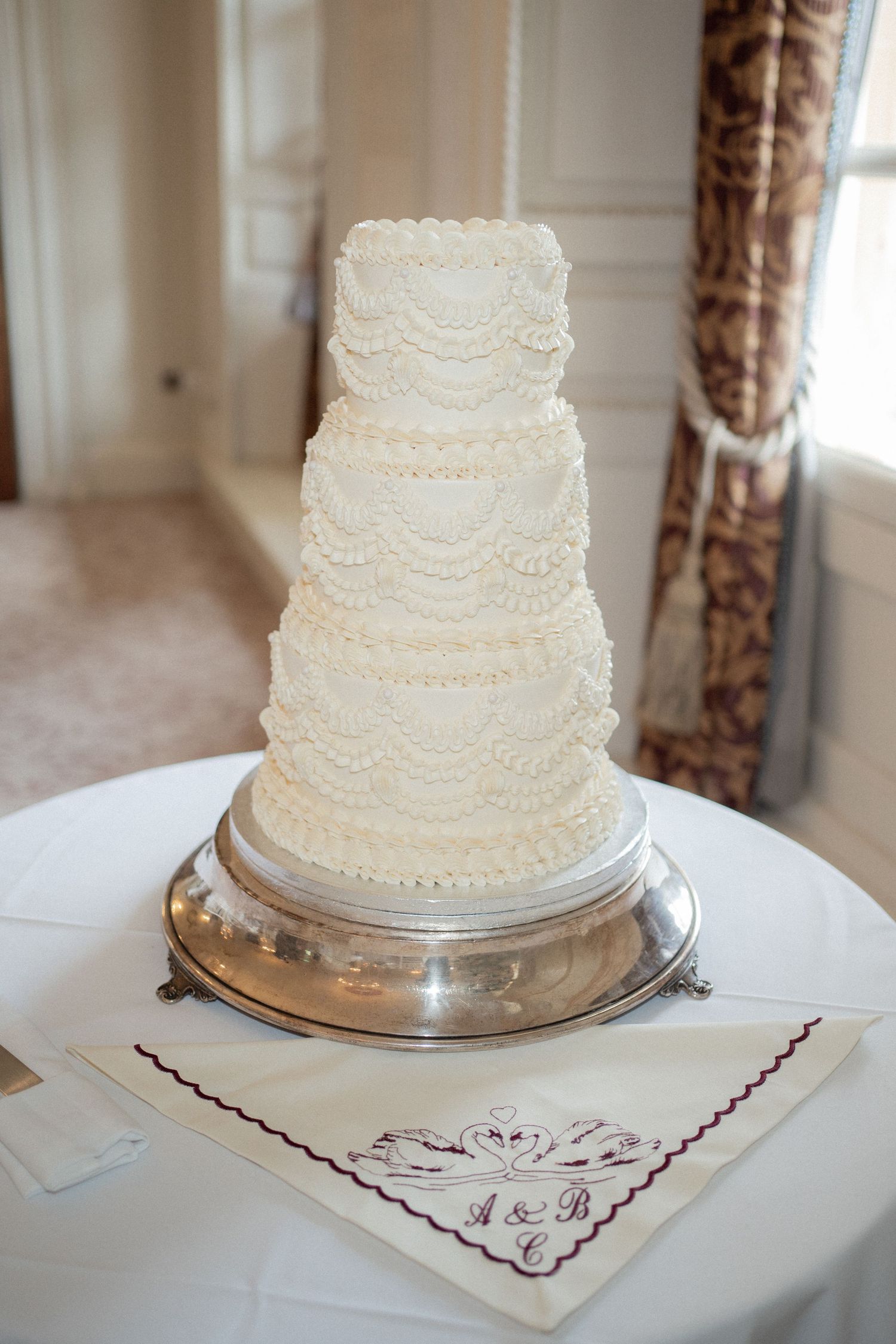 White tiered wedding cake with delicate lace piping details displayed on an embroidered cloth.
