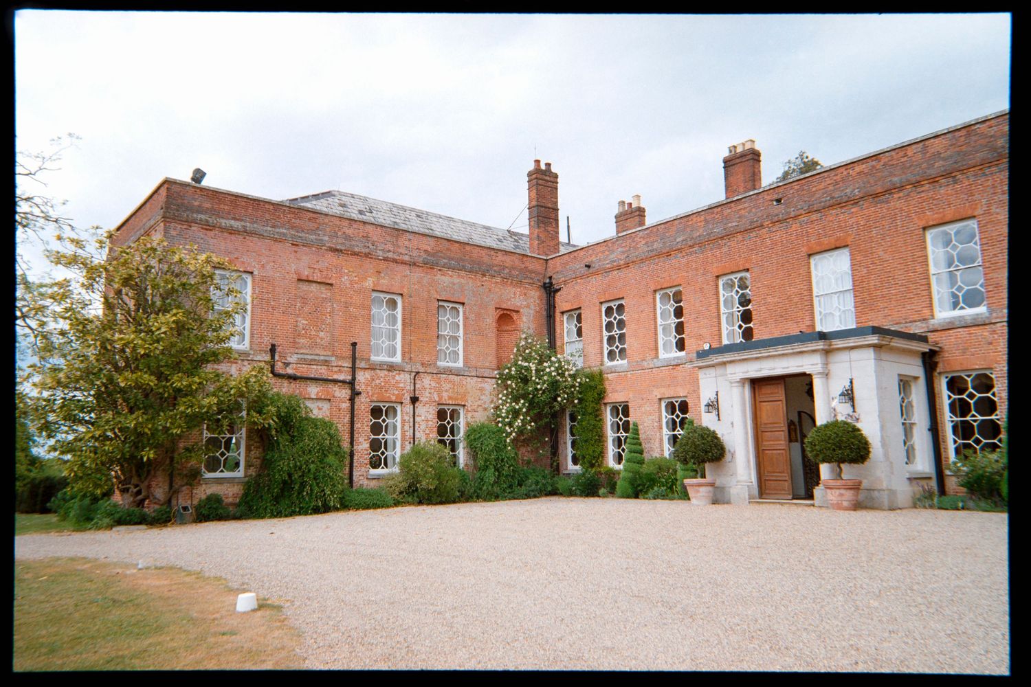 Red brick English country manor with white-framed windows, a stone porch, topiary pots and a gravel drive, ideal for a wedding venue.