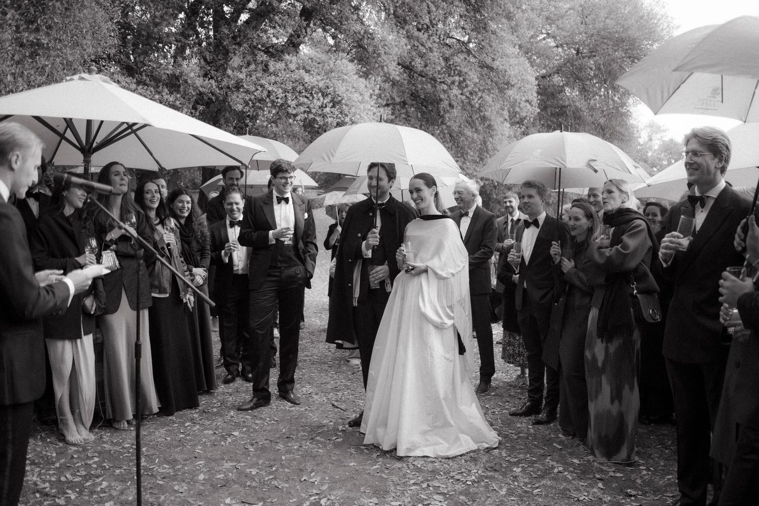 A bride in white and a groom in black walk past guests holding umbrellas in the rain during an outdoor wedding celebration.
