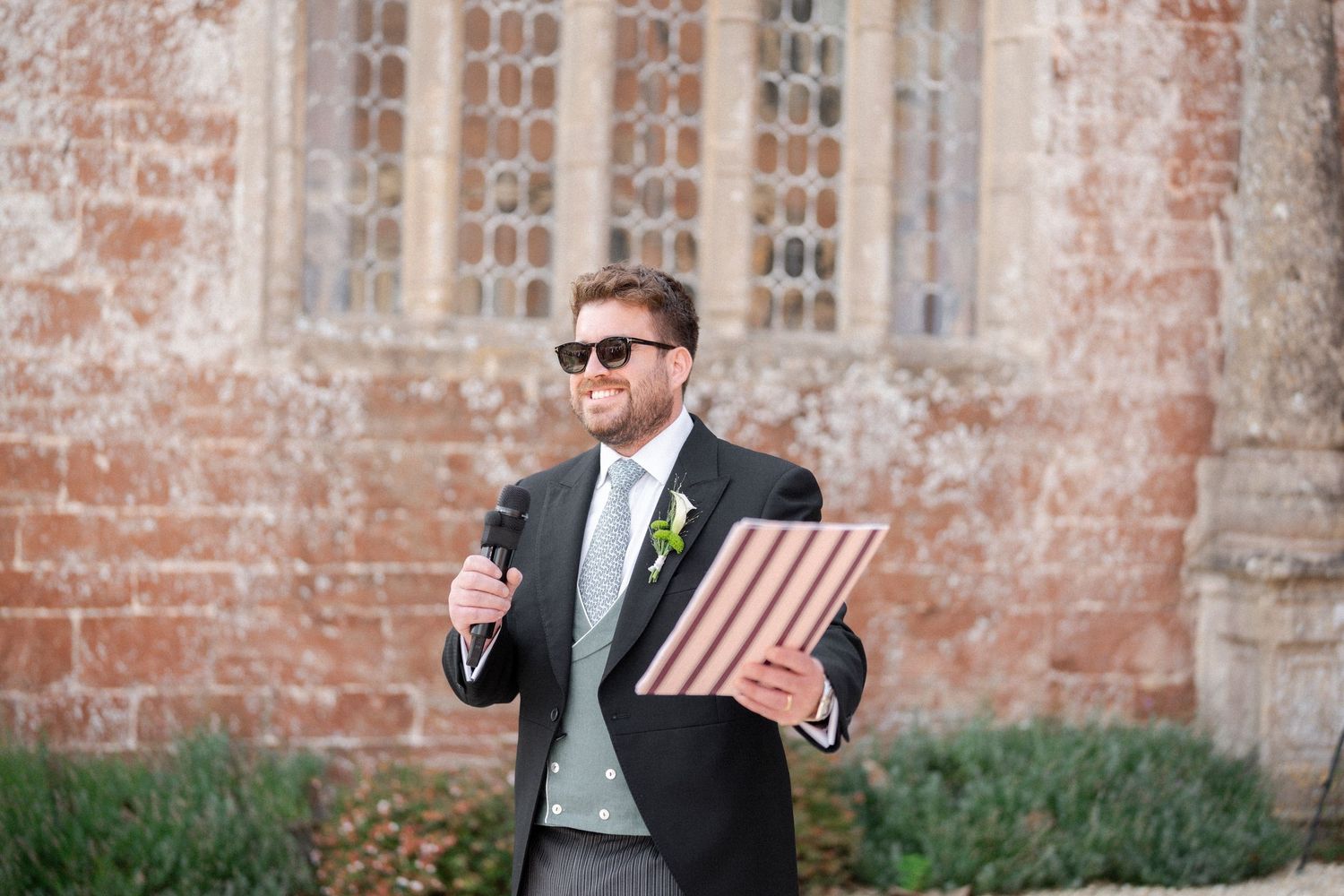 Smiling groom in morning suit and sunglasses holds a striped folder and microphone outside a brick church.