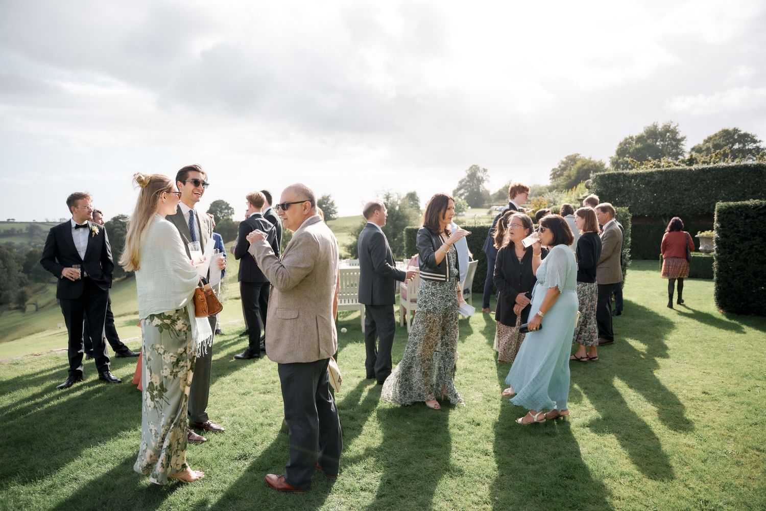 Wedding guests mingle and socialize on a sunny lawn during an outdoor reception celebration.