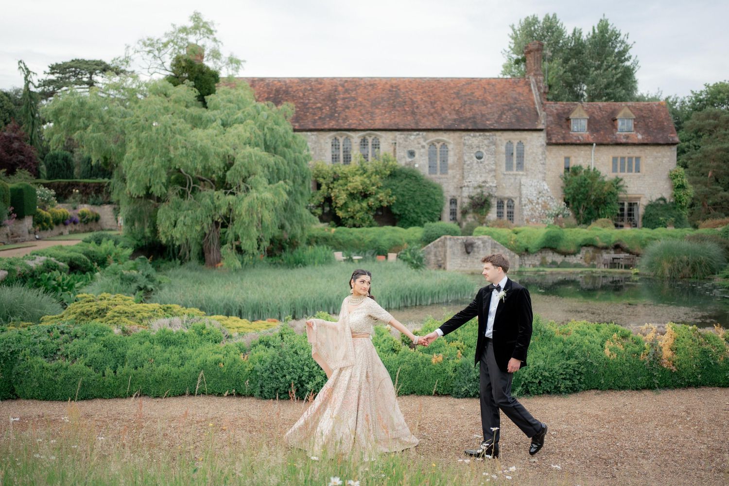 Couple walking hand in hand in front of a historic stone manor house with willow trees.
