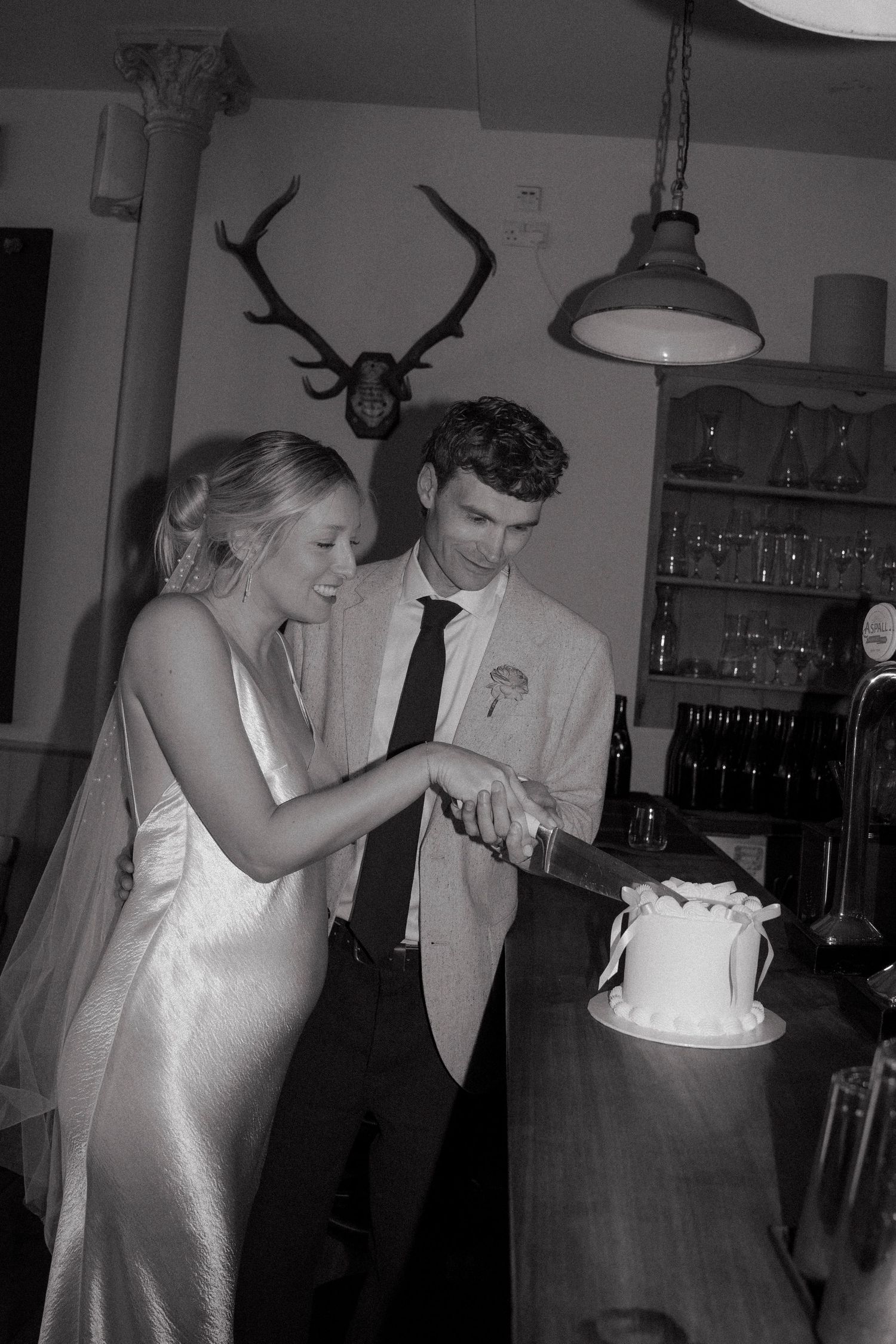 Black and white photo of bride and groom cutting wedding cake together at rustic venue with antler decor.