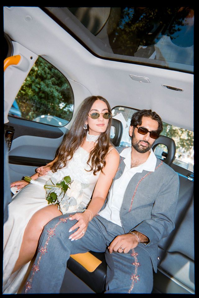 A couple in formal attire sits in a luxury car, woman in white dress holding flowers, man in grey suit, both wearing sunglasses.