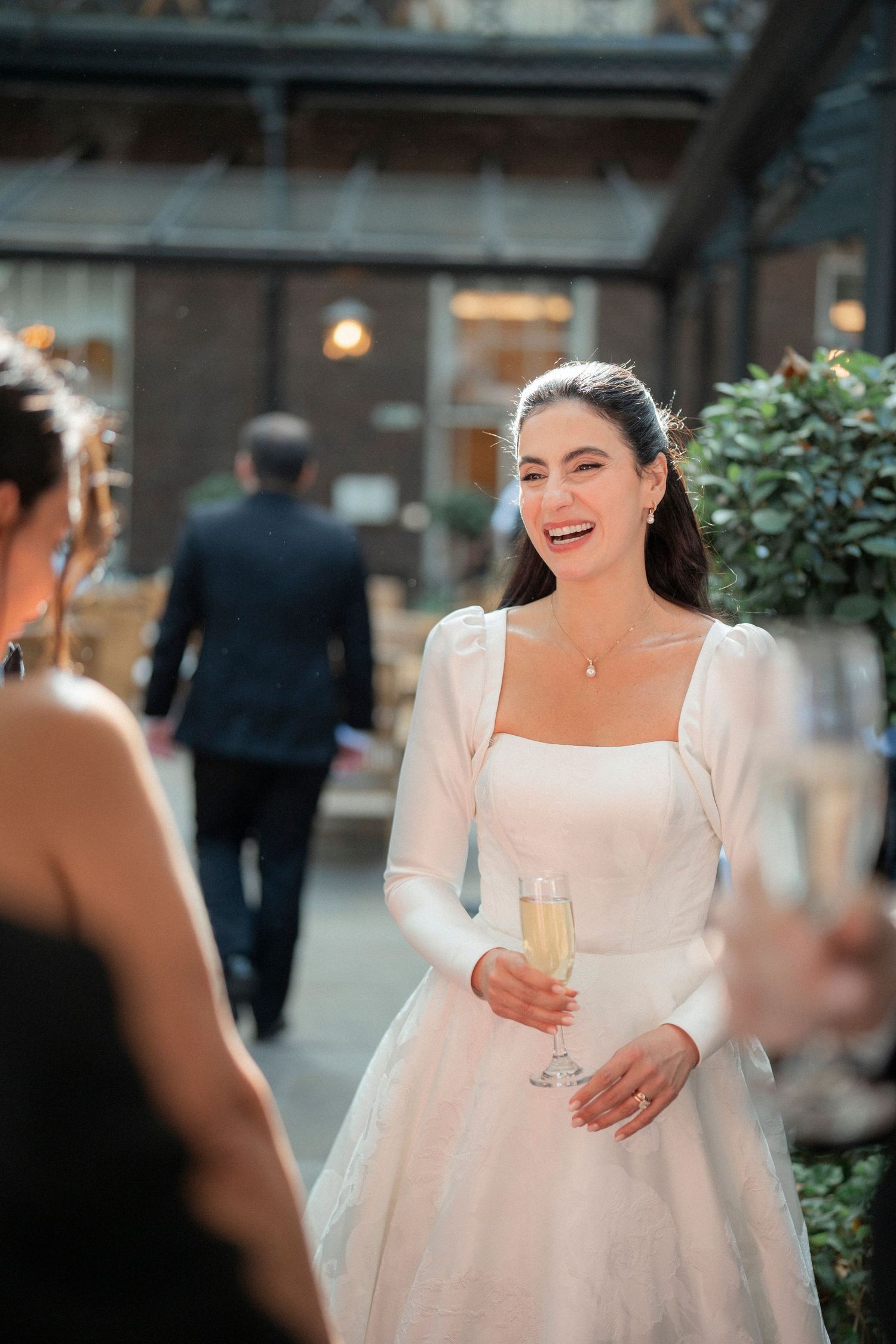 Radiant bride in white gown laughs while holding champagne glass at outdoor wedding reception.