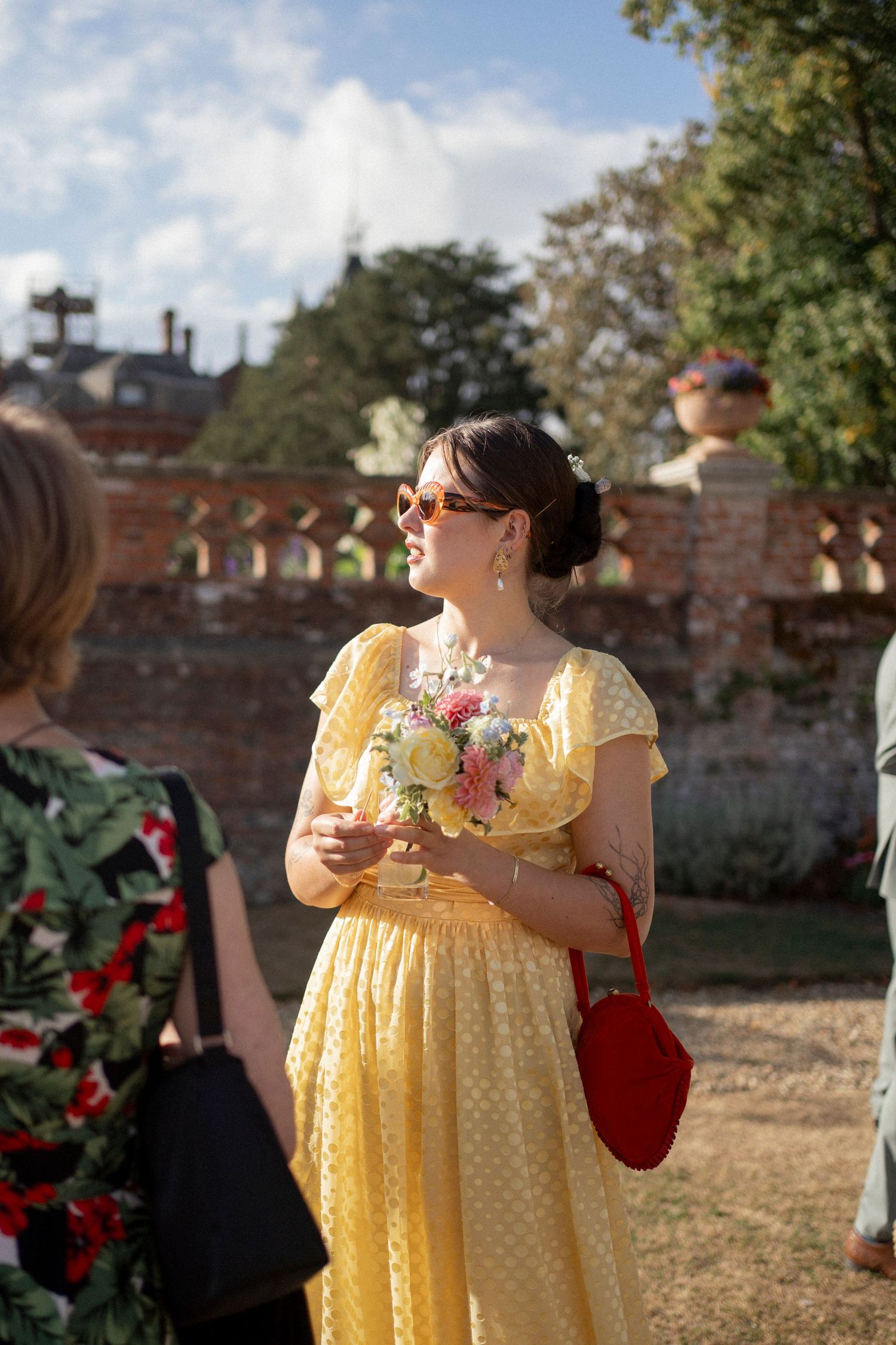 Woman in yellow dress holding floral bouquet at outdoor garden wedding venue on sunny day.