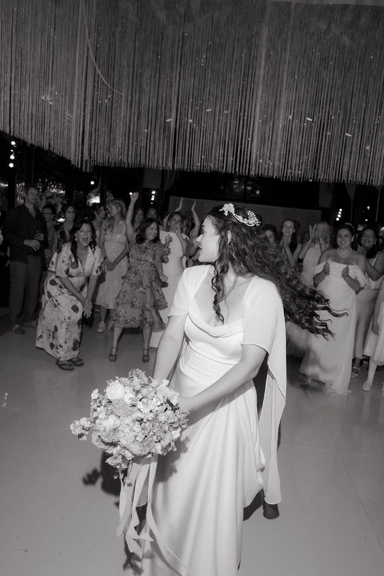 Bride in white gown prepares to toss bouquet to excited guests at wedding reception in black and white photo.