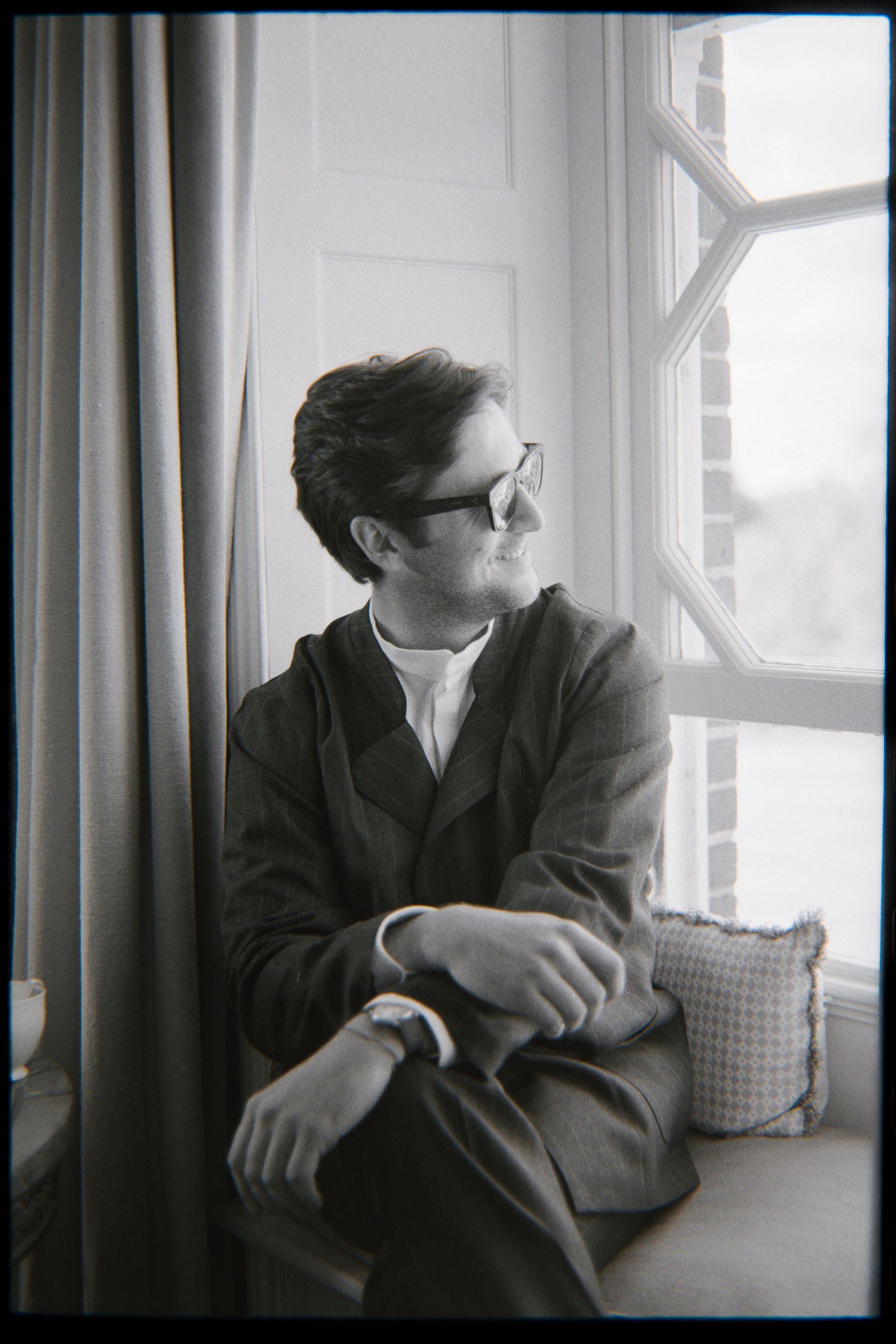 Man in dark glasses and a wool jacket sits on a cushioned window seat, gazing outside in black and white.
