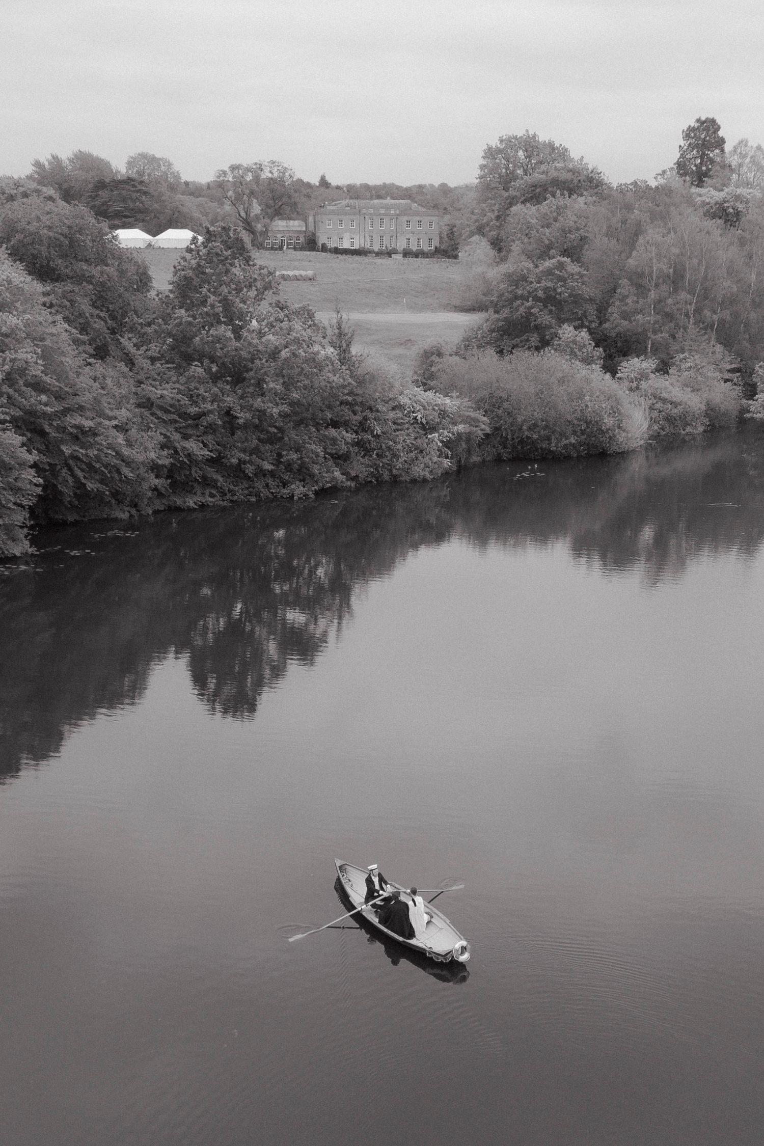 Black and white photo of a rowing boat gliding on a calm lake, with trees and a stately country house in the background.