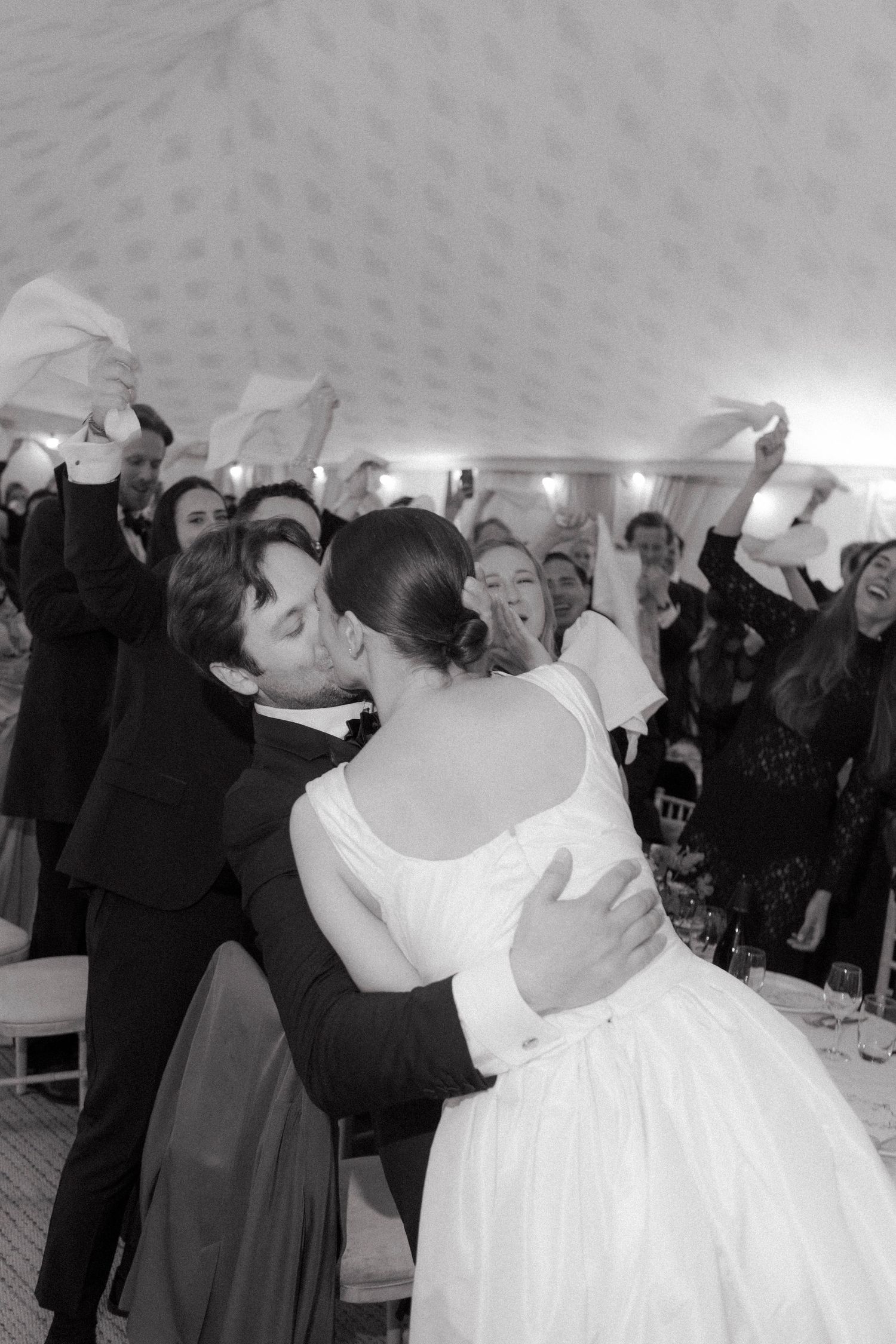 Black and white photo of a bride in a full-skirted gown embracing a groom in a tuxedo while guests wave napkins in a tented reception.