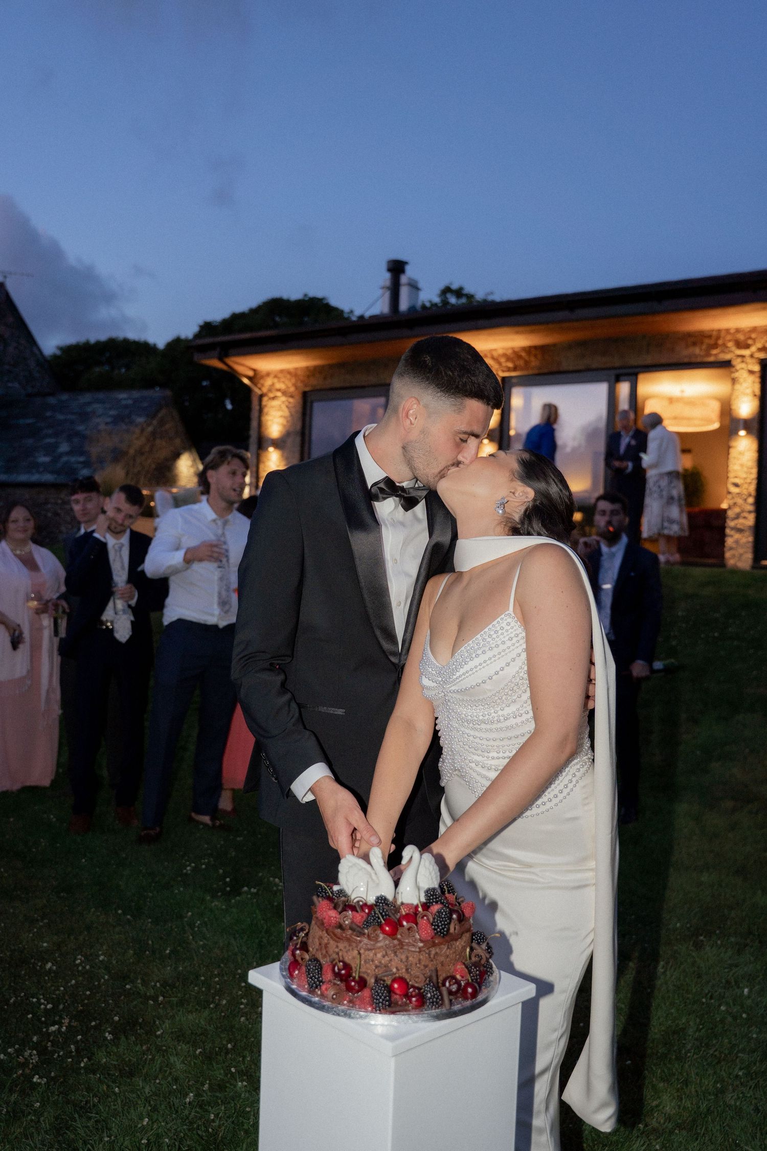 Newlyweds cut their wedding cake together at an outdoor evening reception.