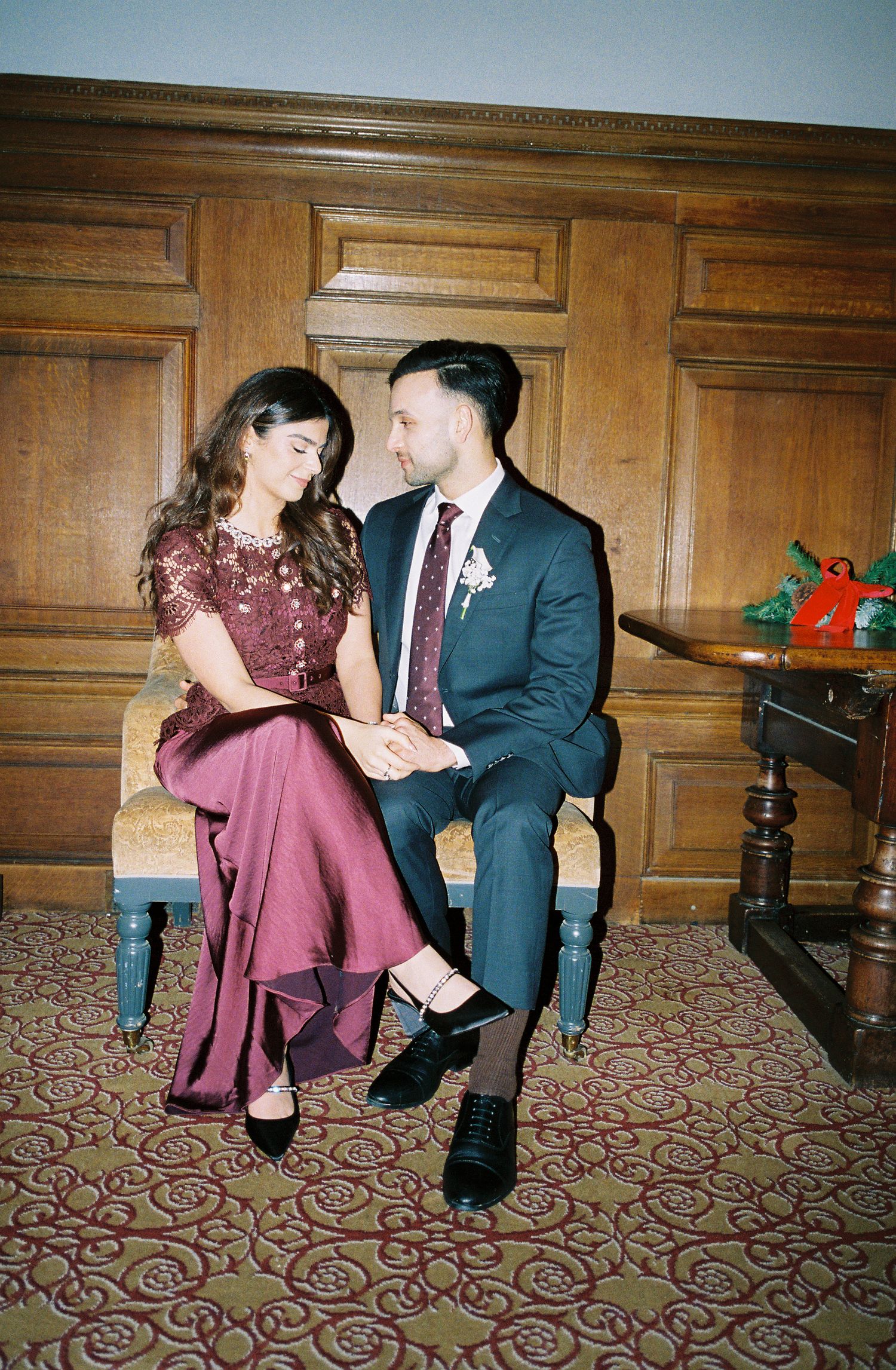 Couple in formal attire sitting together, woman in burgundy lace gown, man in dark suit with floral boutonniere, elegant wood-paneled room.