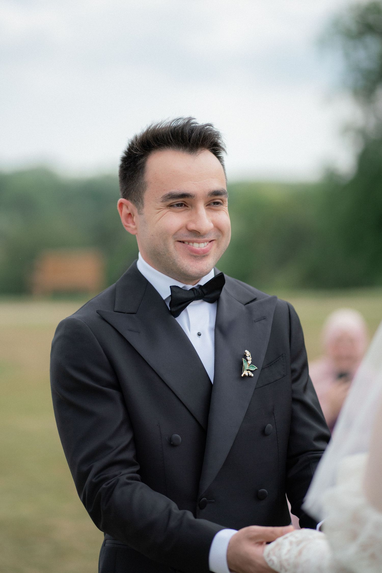 A groom in a black tuxedo smiles during an outdoor wedding ceremony.
