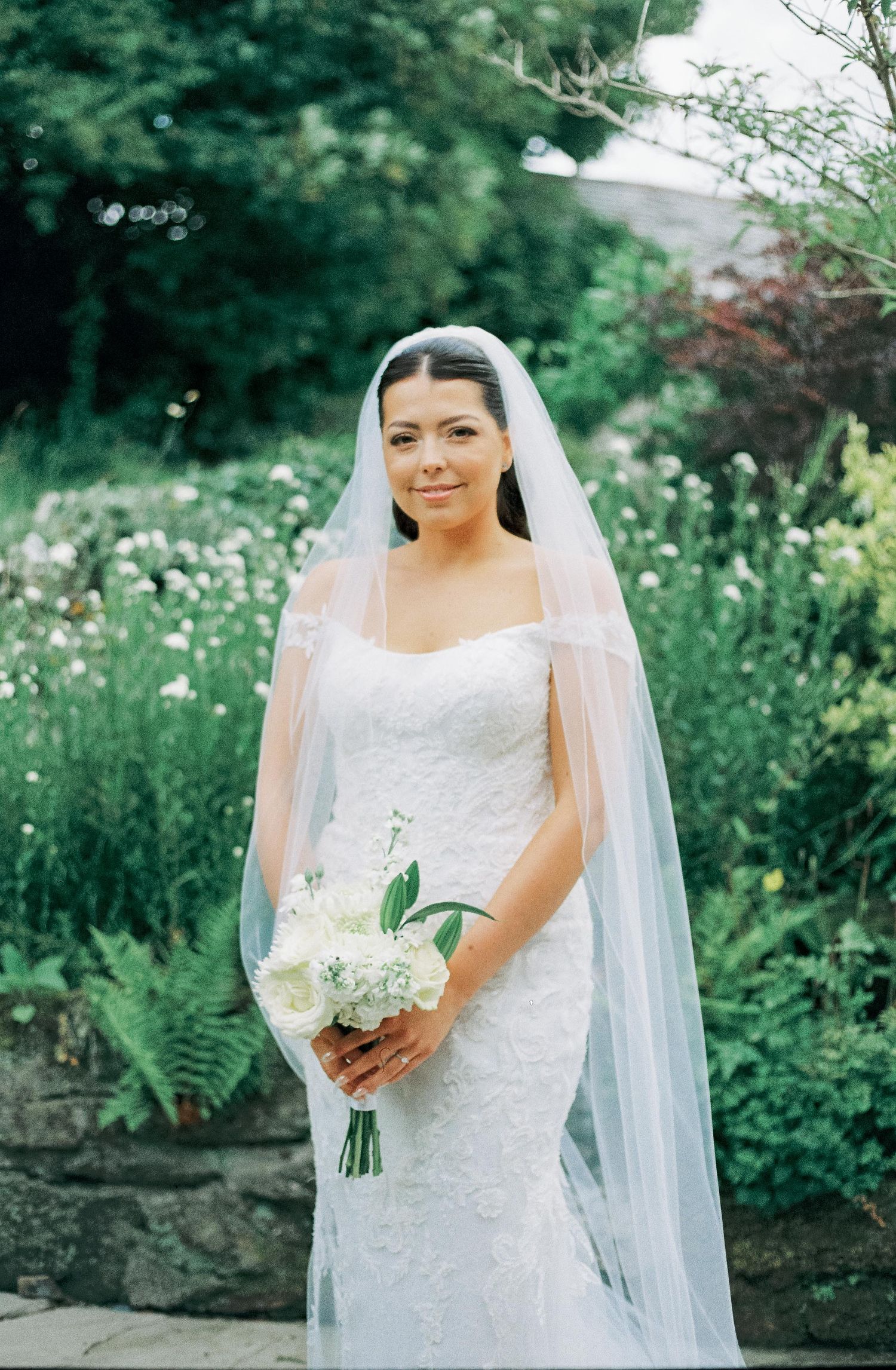 A bride in a classic white lace wedding dress and veil poses in a lush garden setting.