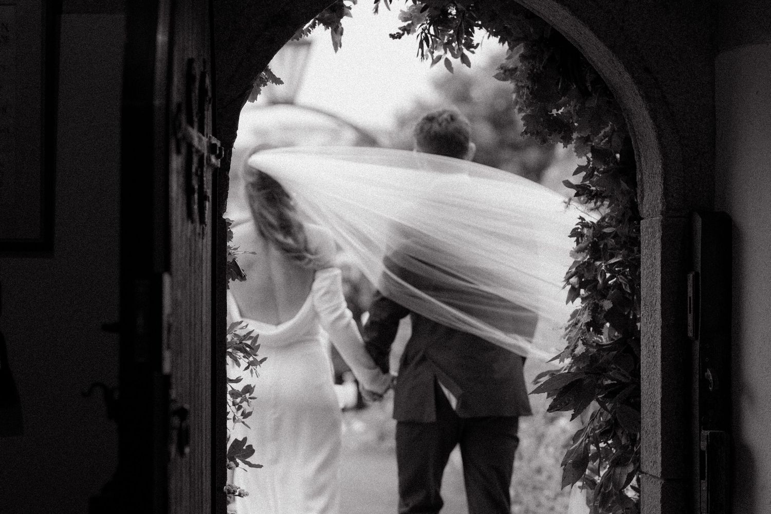 Black and white wedding photograph showing a bride's veil flowing in the wind against a dark architectural background.