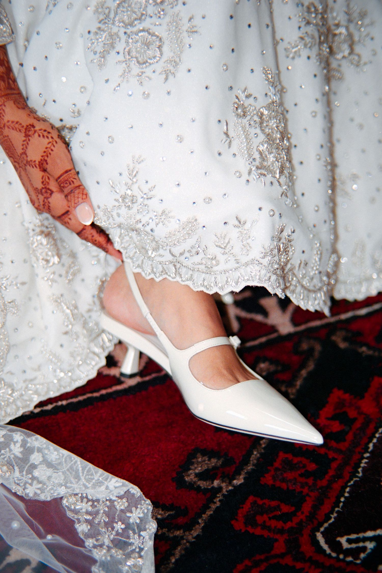 Close-up view of a white bridal shoe with lace dress hem against a red and black patterned carpet.