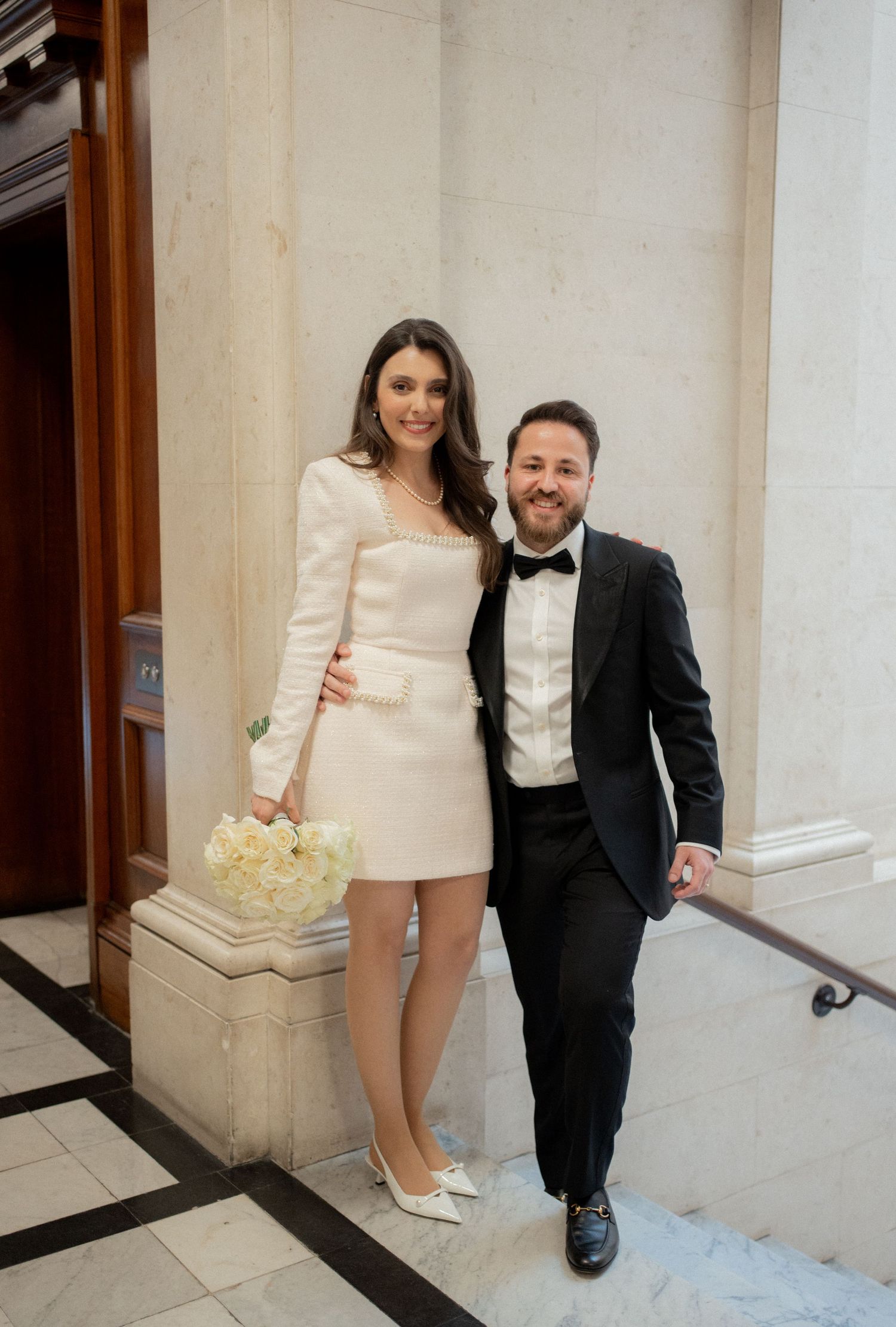 A couple poses together at their wedding, with the bride in a short white dress and the groom in a classic black tuxedo.