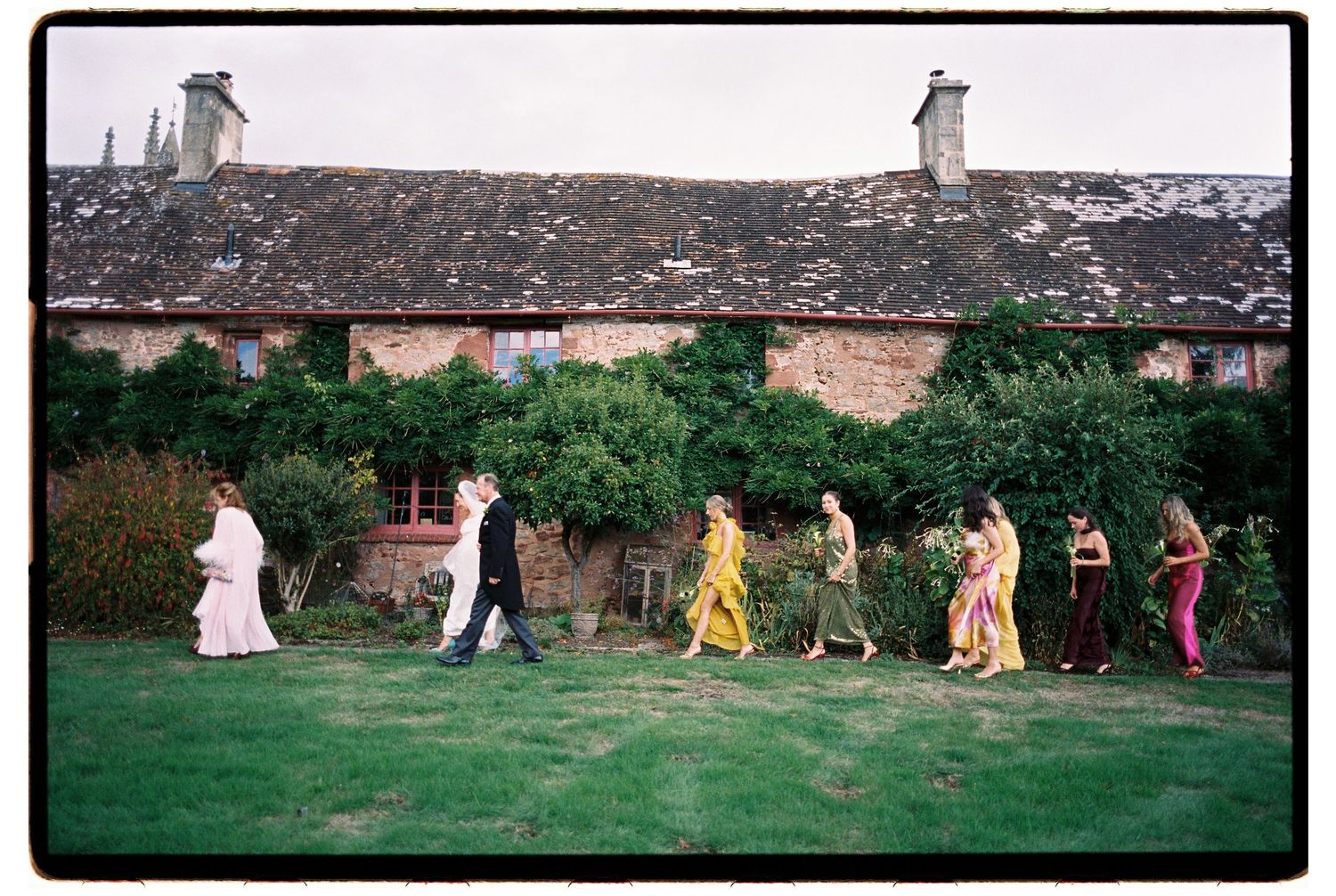 Wedding party walking across green lawn in front of rustic stone cottage with moss-covered roof.