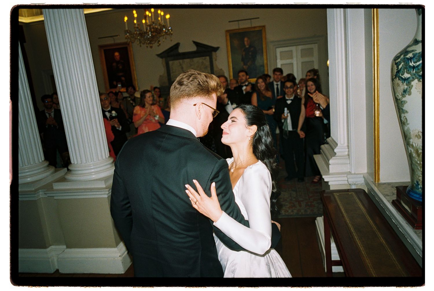 Bride in a white gown and groom in a dark suit share a first dance in an elegant ballroom as wedding guests watch beneath a chandelier.