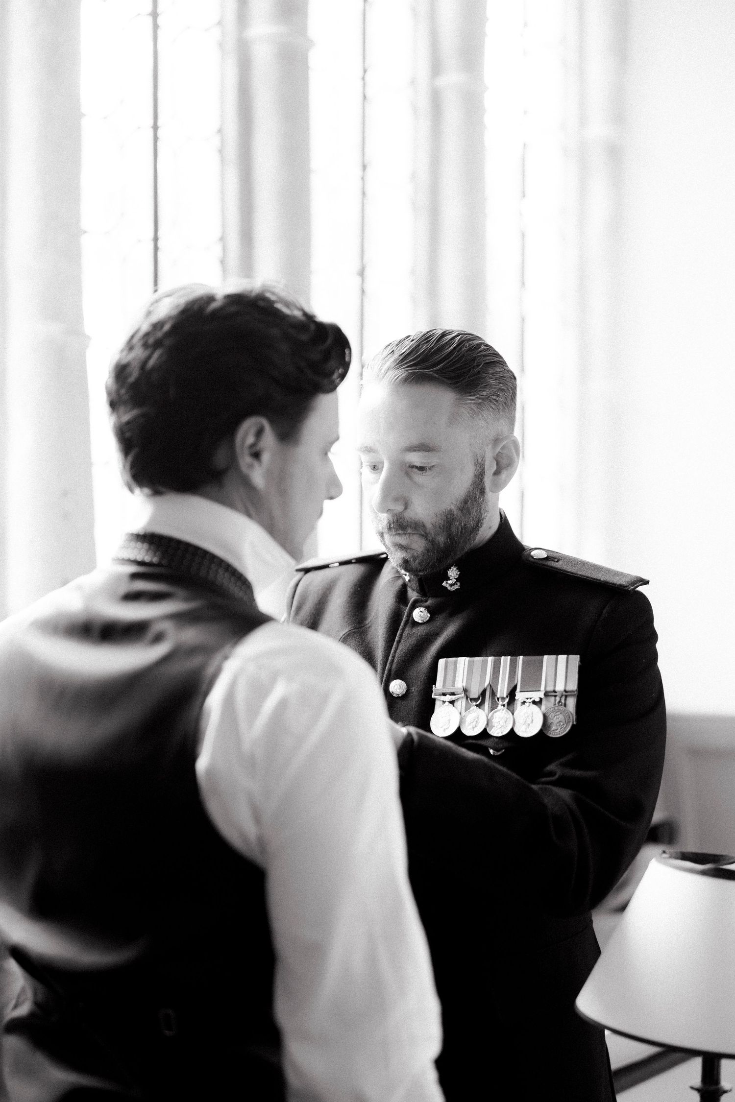 Groom in military dress uniform with medals being assisted by groomsman near window in black and white.