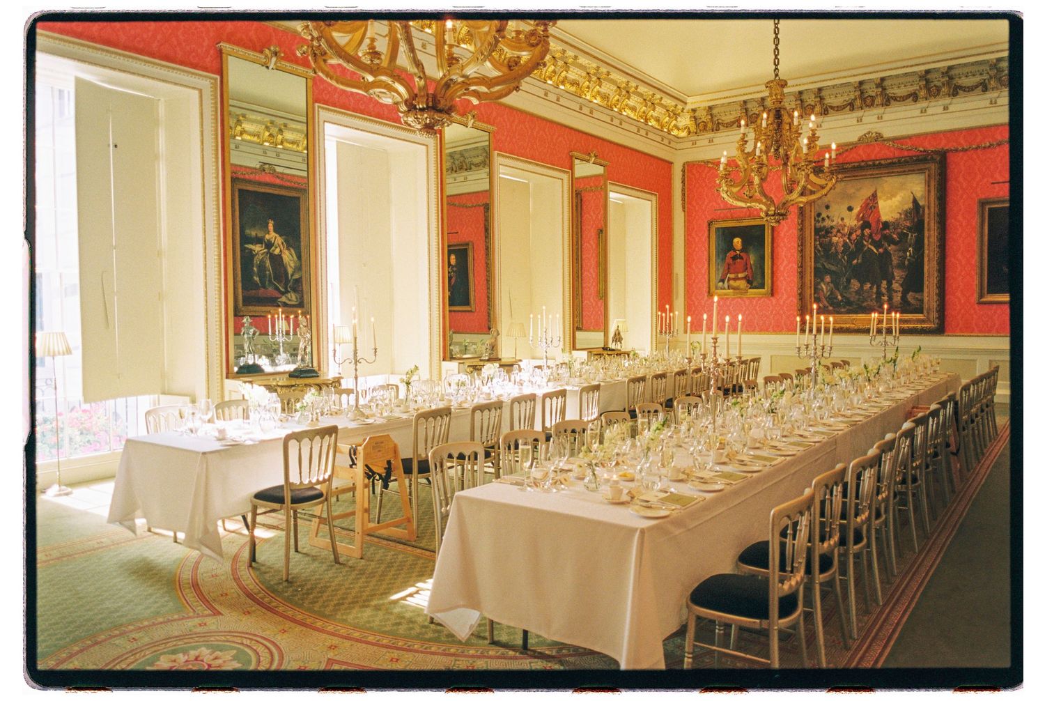 Elegant wedding reception room with long white tables, gold chandeliers, and red walls adorned with classic paintings.