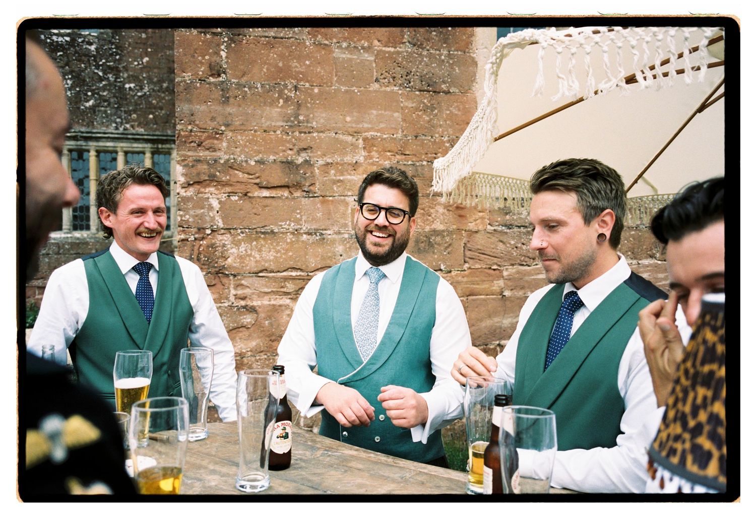Groomsmen in green vests and ties laughing together outdoors at a wedding reception with drinks on table.