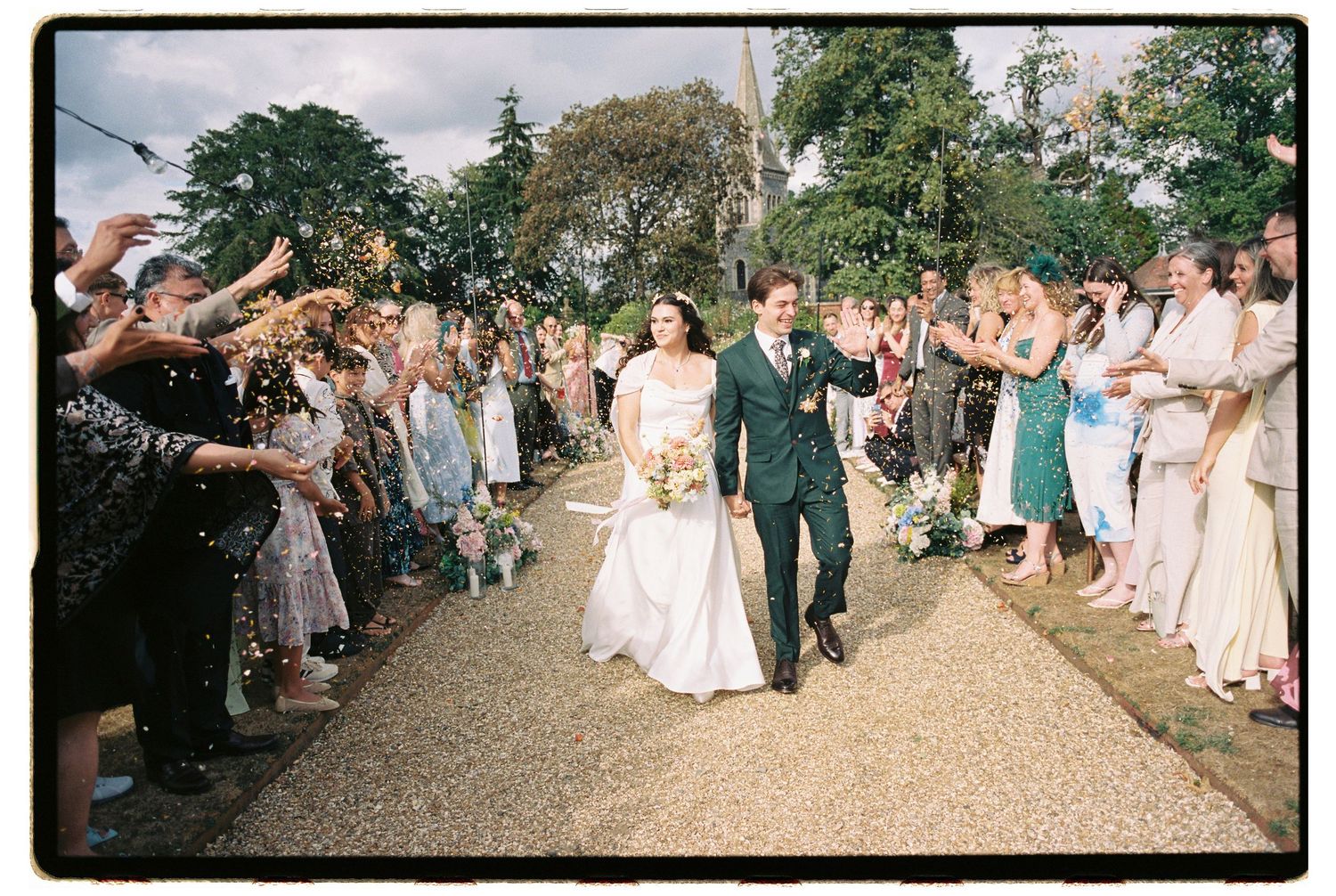 Bride and groom walk through confetti shower with cheering guests lining path at outdoor wedding celebration.