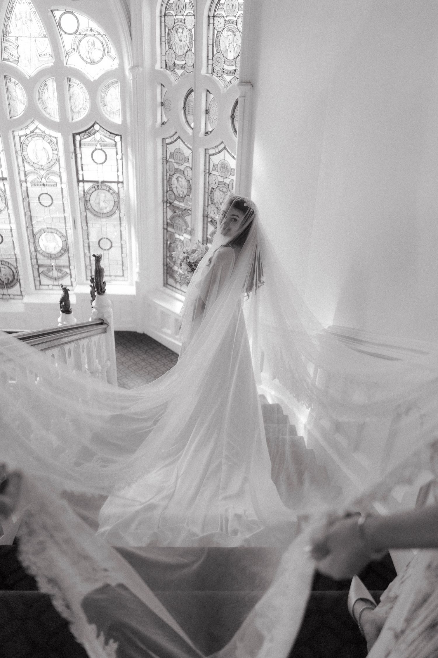 Black and white photo of a bride in a flowing gown and long veil on a grand staircase with ornate stained glass windows.
