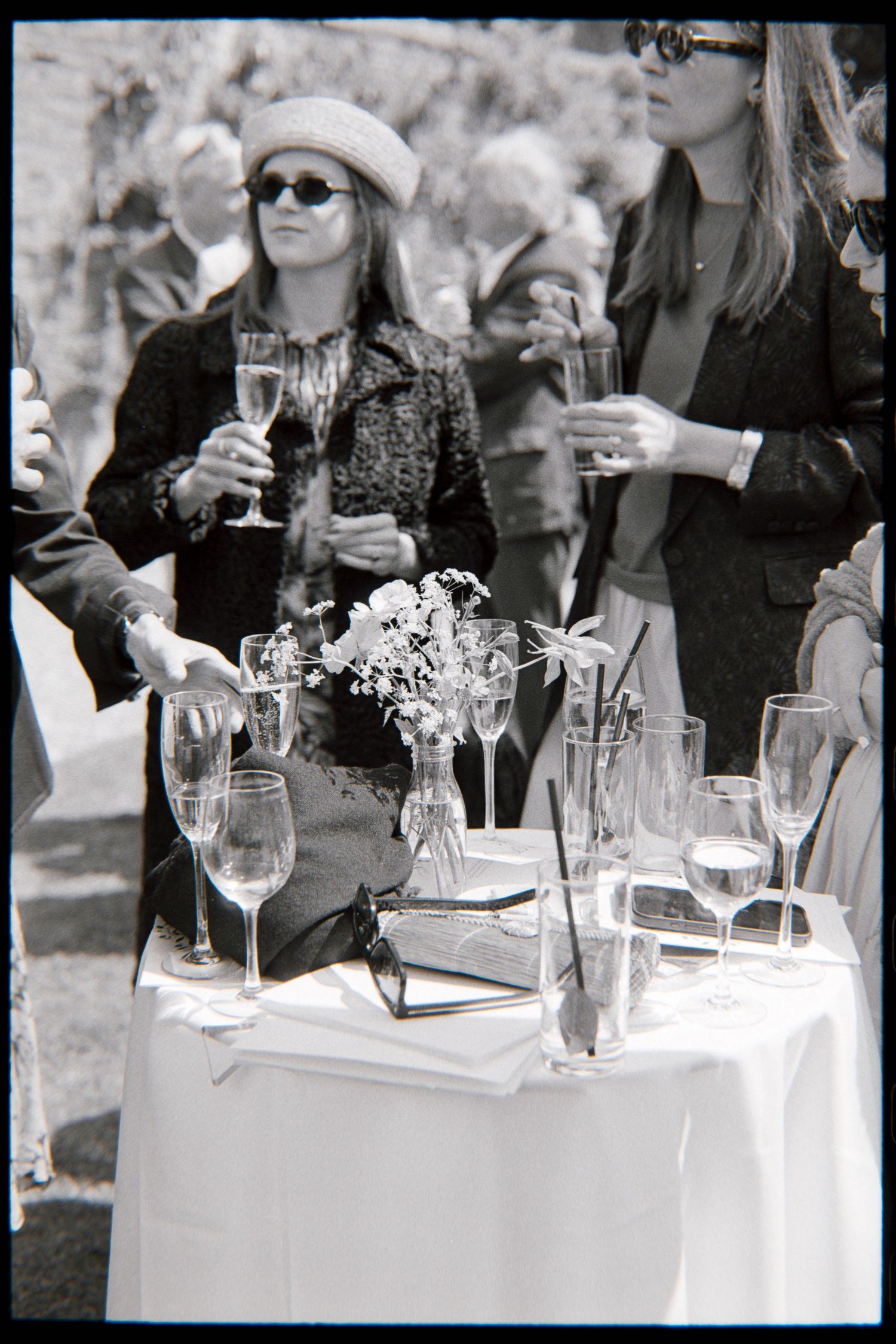 Black and white photo of wedding guests in sunglasses holding champagne glasses beside a table with flowers and drinks.