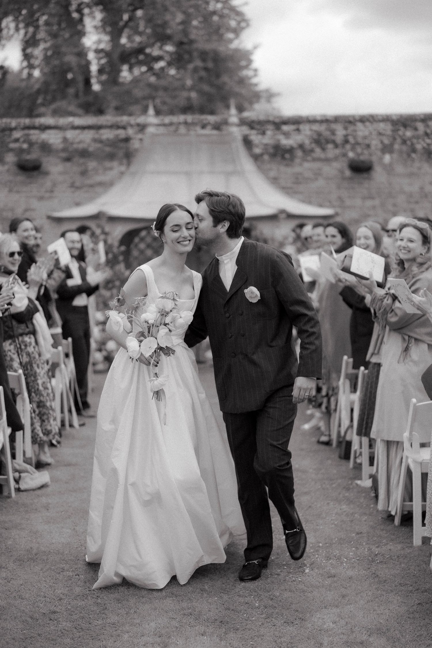 Bride in a flowing gown holds a bouquet as the groom kisses her cheek while guests applaud along the aisle outdoors.