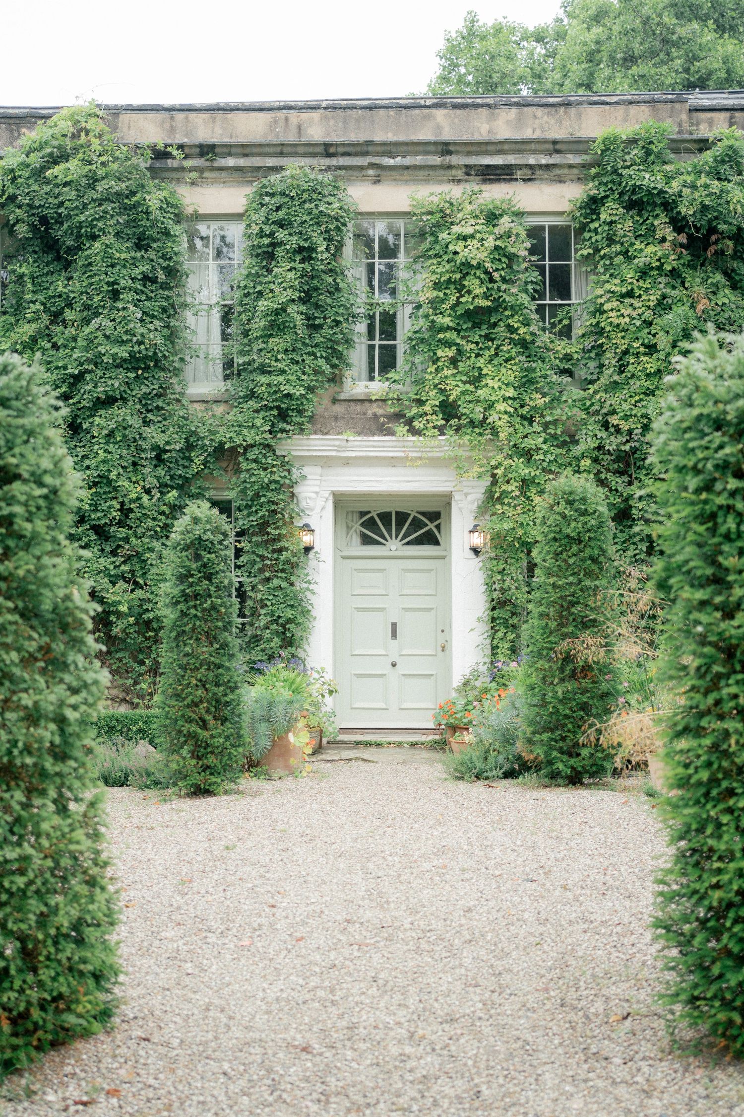 Ivy-covered stone manor house with a mint green door, flanked by manicured topiary hedges along a gravel pathway.