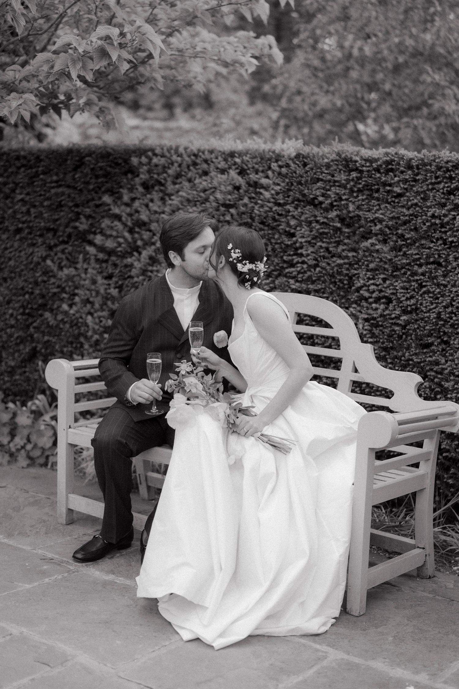 Black and white photo of a bride and groom kissing on a wooden garden bench, holding champagne flutes and a bouquet.